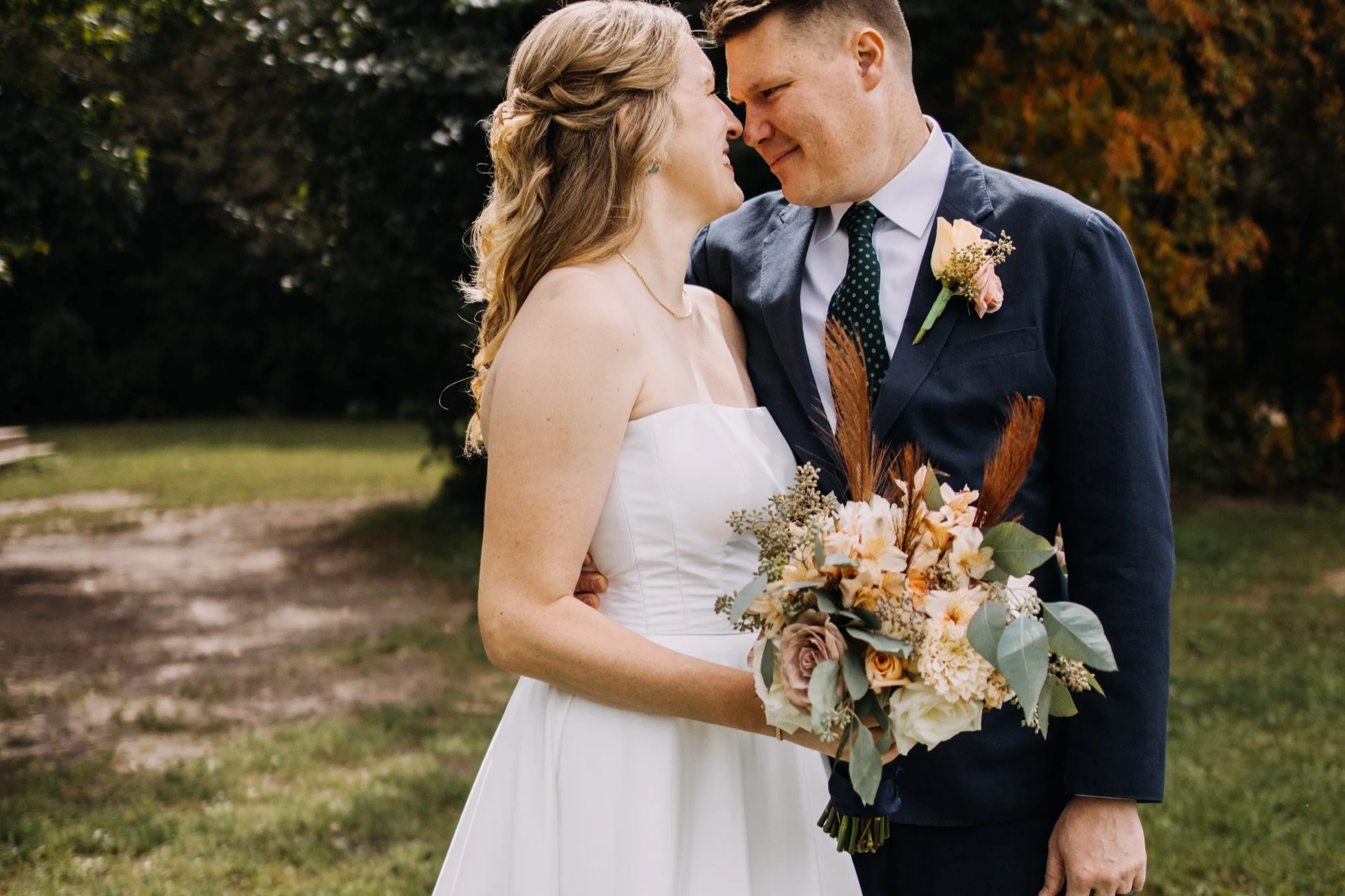 A bride and groom stand close together outdoors, smiling and touching foreheads. The bride is in a white strapless wedding dress holding a bouquet of flowers, and the groom is in a dark suit with a patterned tie and a boutonniere. The background includes greenery and trees.