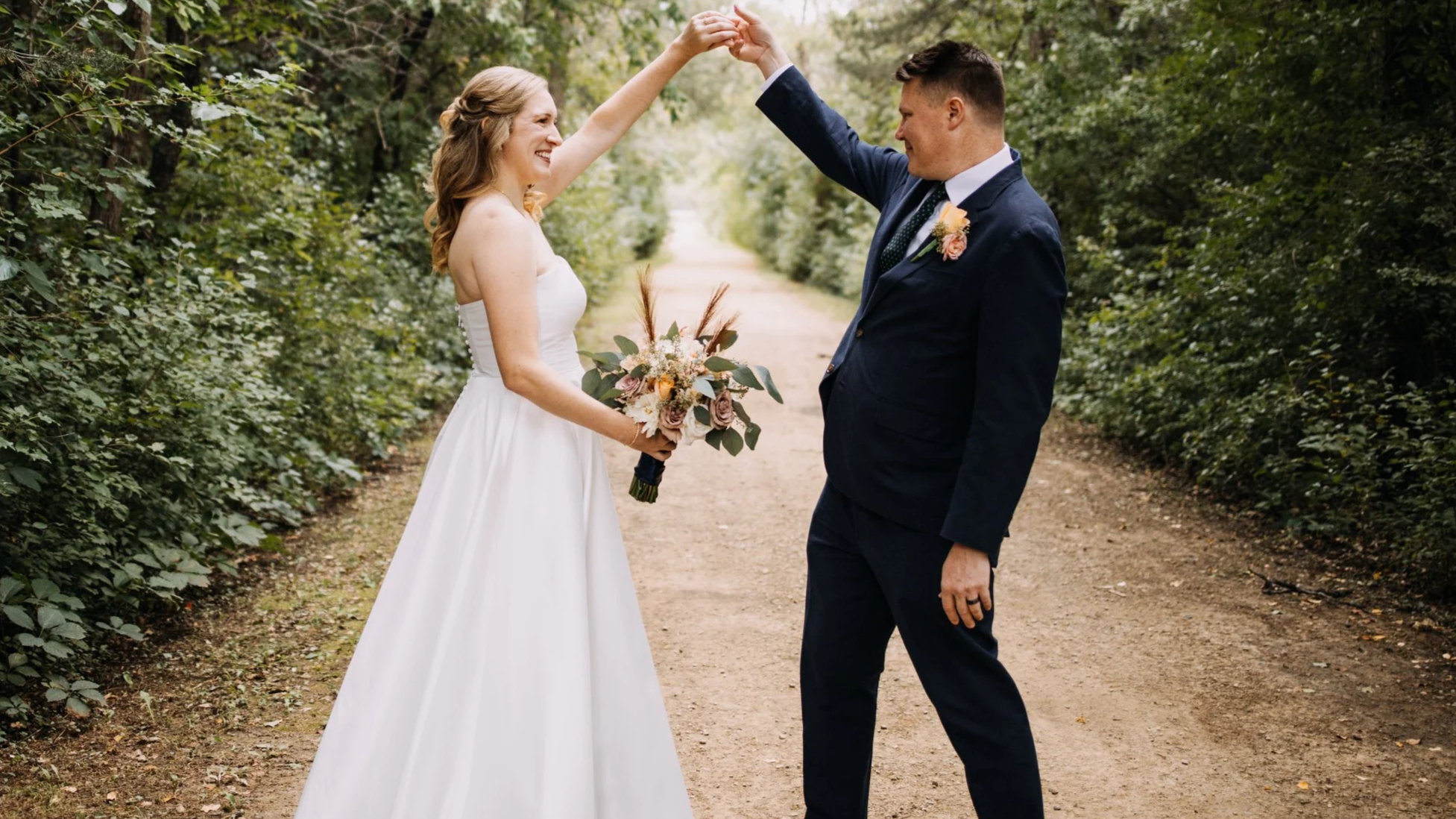 A bride and groom dancing outdoors on a dirt path surrounded by green foliage, with the bride holding a bouquet and both smiling.