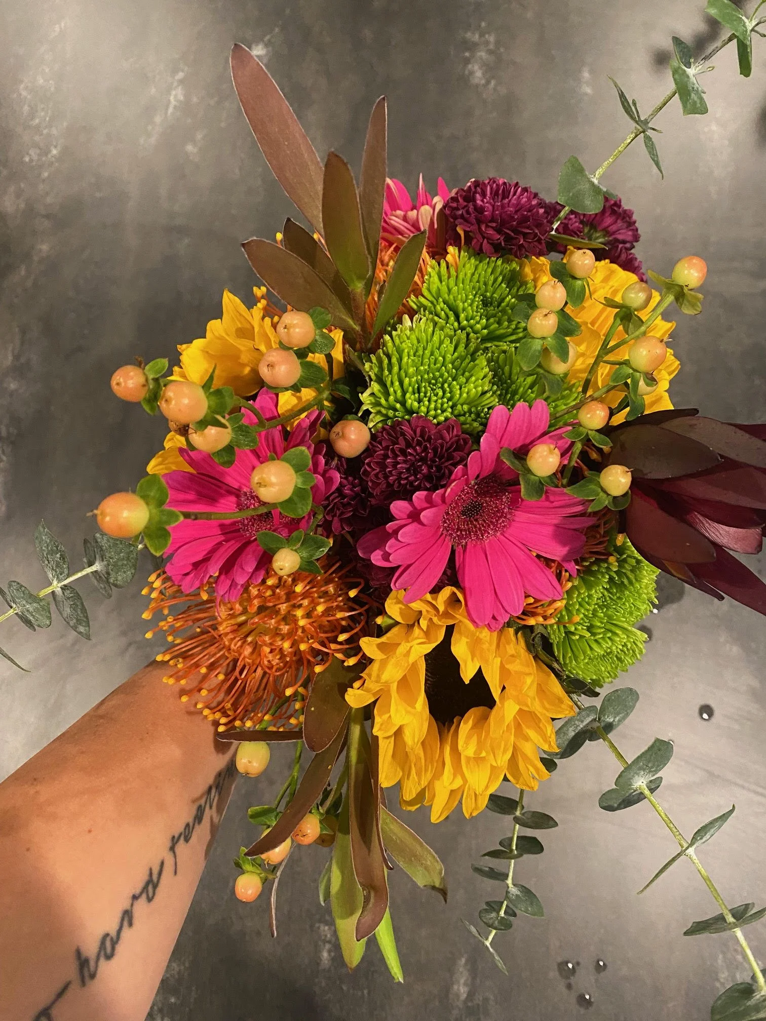 Colorful bouquet of pink, yellow, green, and purple flowers with green and brown leaves being held by a hand with a tattoo.