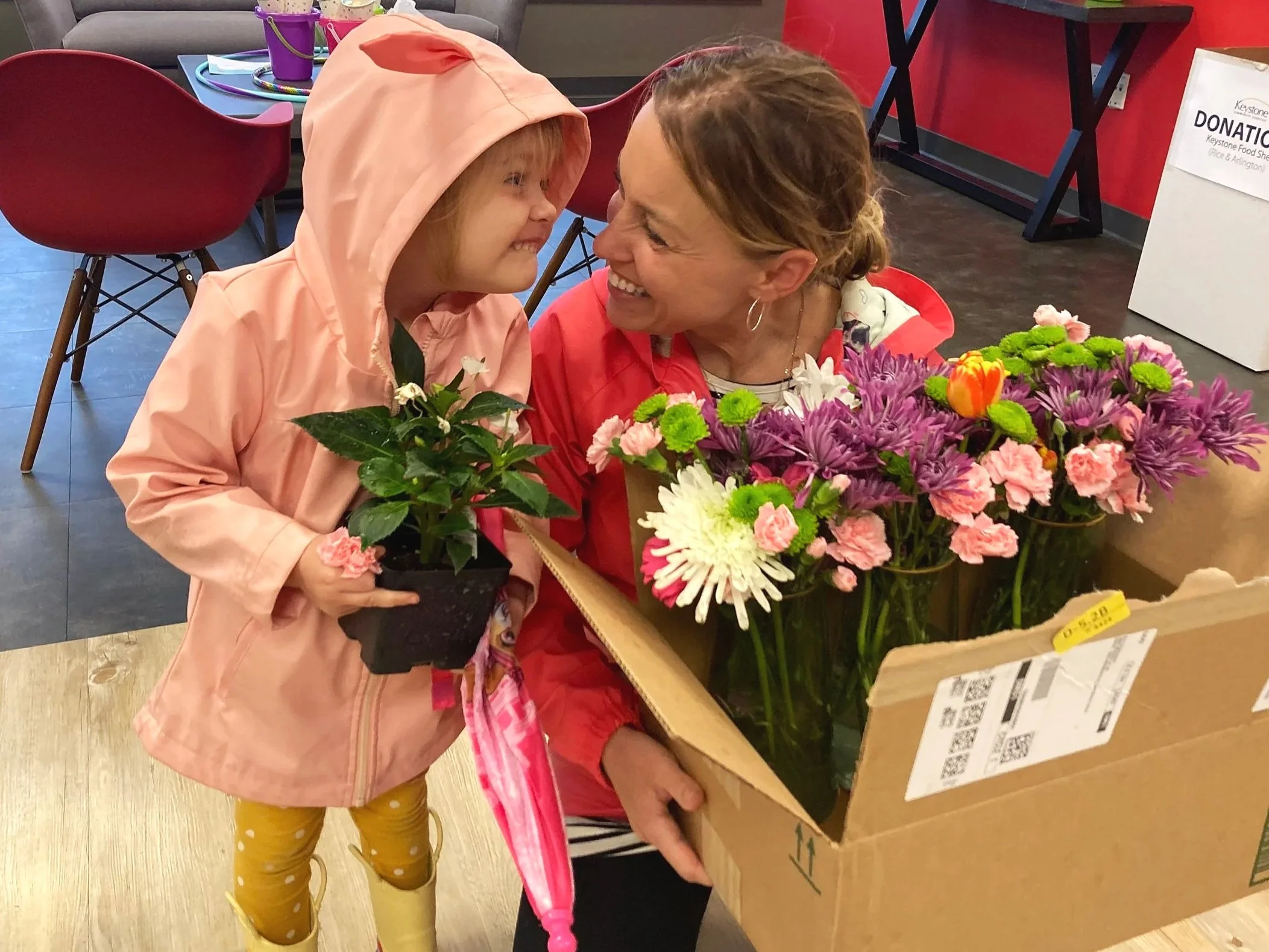 A young girl in a pink hooded raincoat and yellow rain boots holding a small potted plant, smiling and leaning close to a woman who is sitting and holding a large box of colorful flowers; they are sharing a joyful moment.