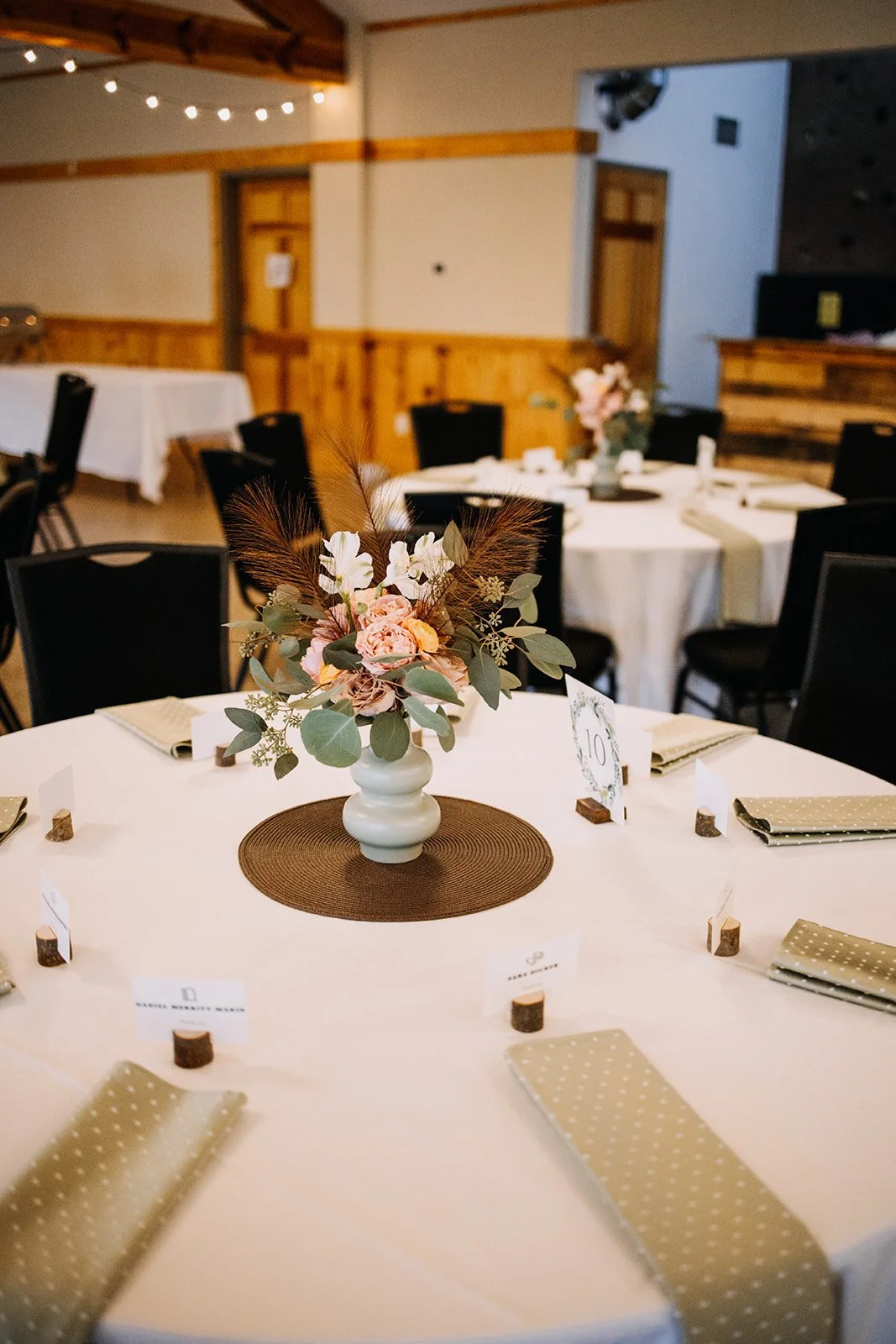 Wedding reception table with a flower centerpiece in a white vase, beige polka dot napkins, and wooden accent decor, in a cozy indoor venue with wood accents and string lights.