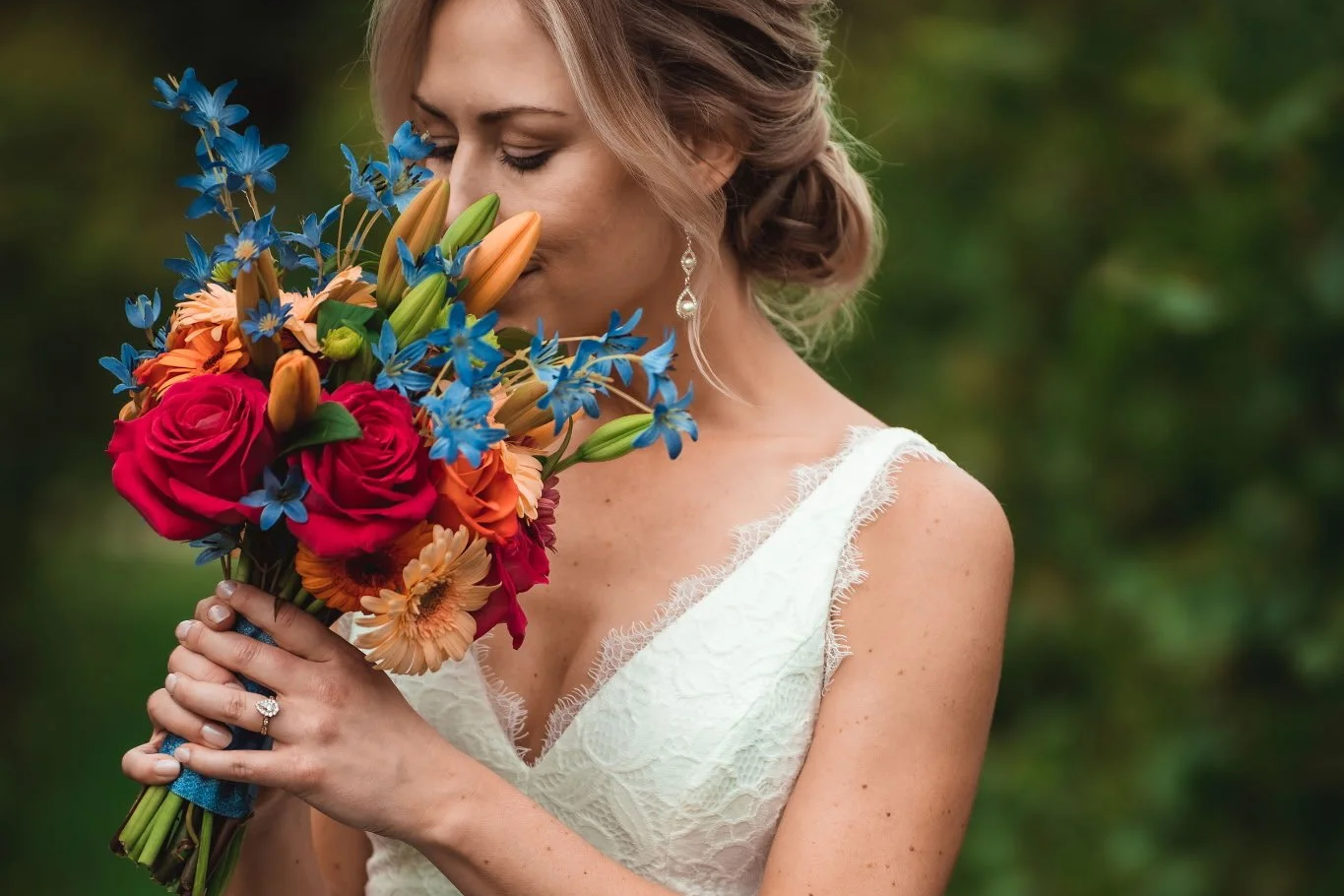 A bride in a white lace dress holding a colorful bouquet of red, orange, blue, and green flowers with her eyes closed, outdoors with blurred green background.