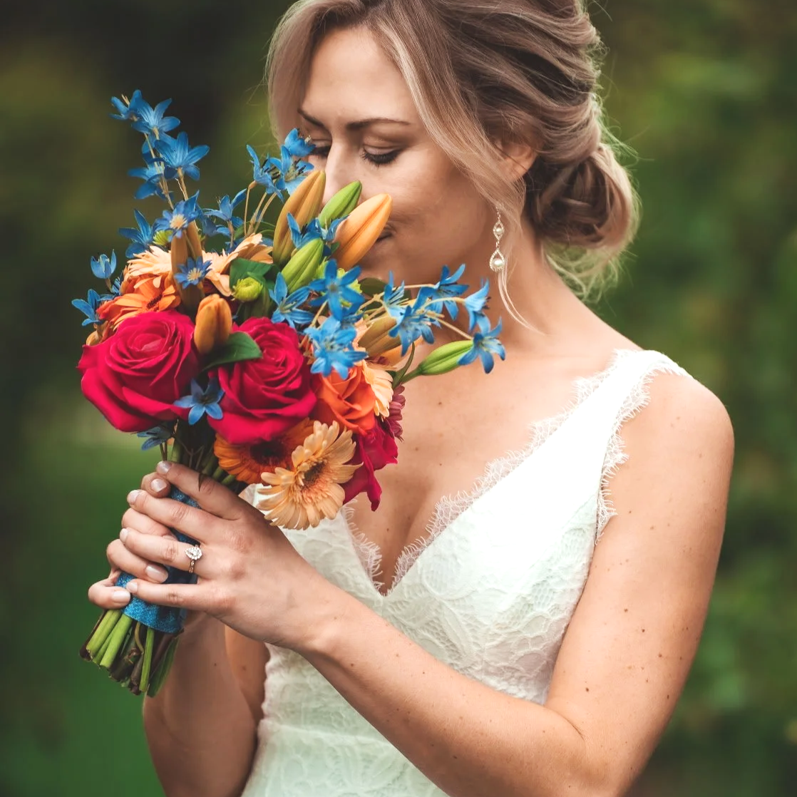 A bride in a white dress holding a colorful bouquet of flowers and smelling it, outdoors with a blurred green background.