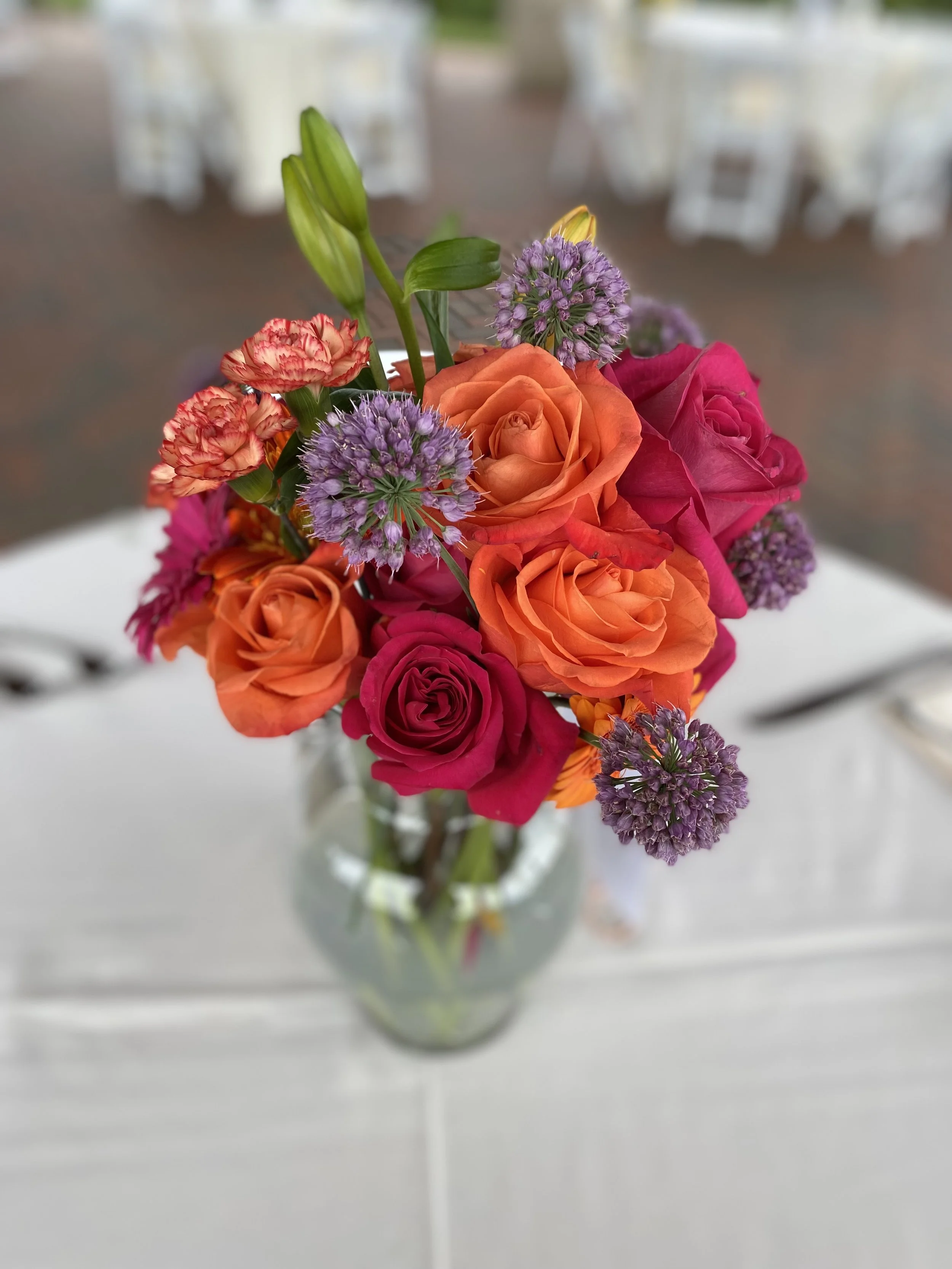 A colorful flower bouquet with roses, purple alliums, and other flowers in a glass vase on a white table.