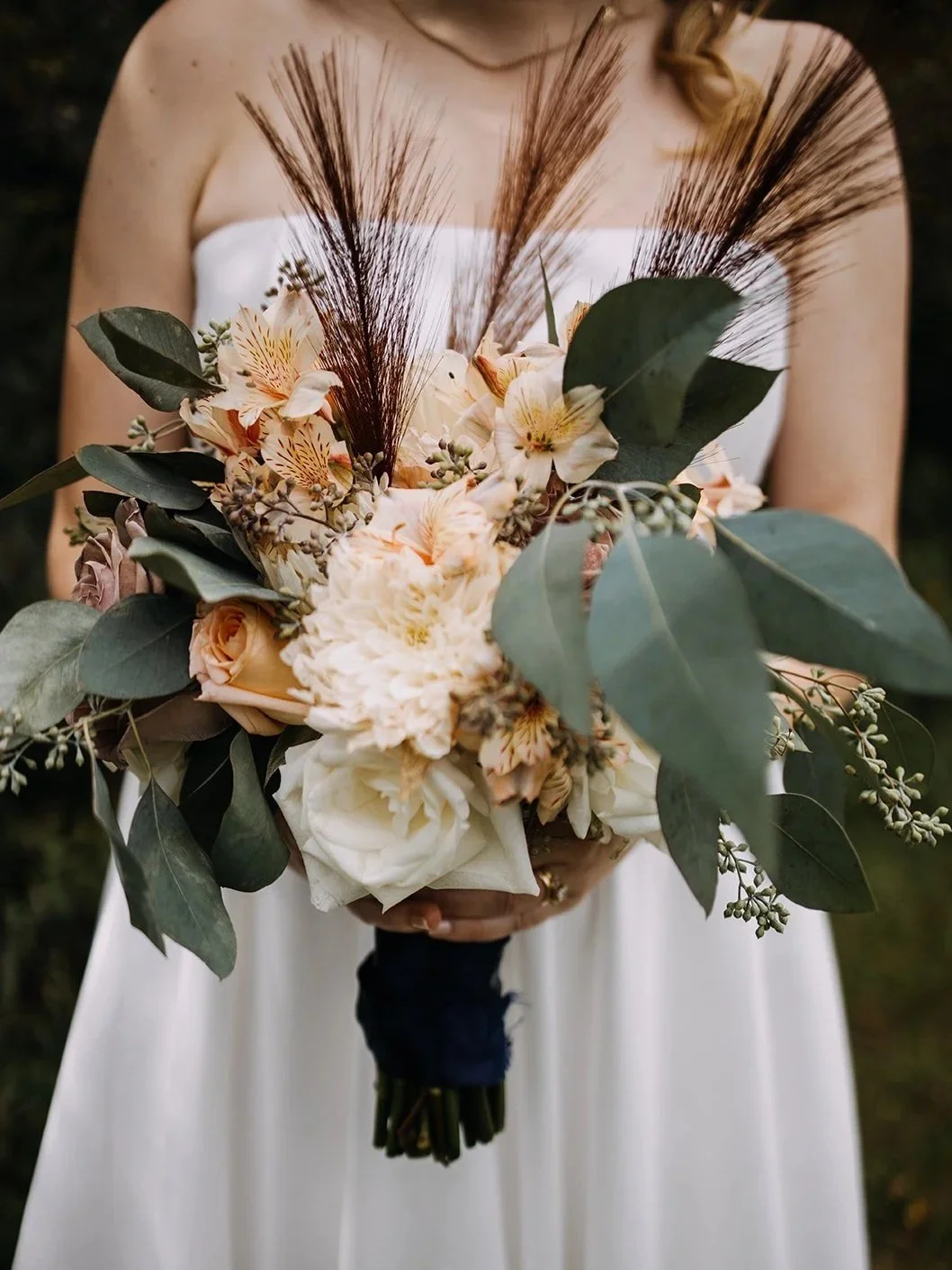 A bride in a strapless white wedding dress holding a bouquet of cream and peach flowers with greenery and decorative feathers.