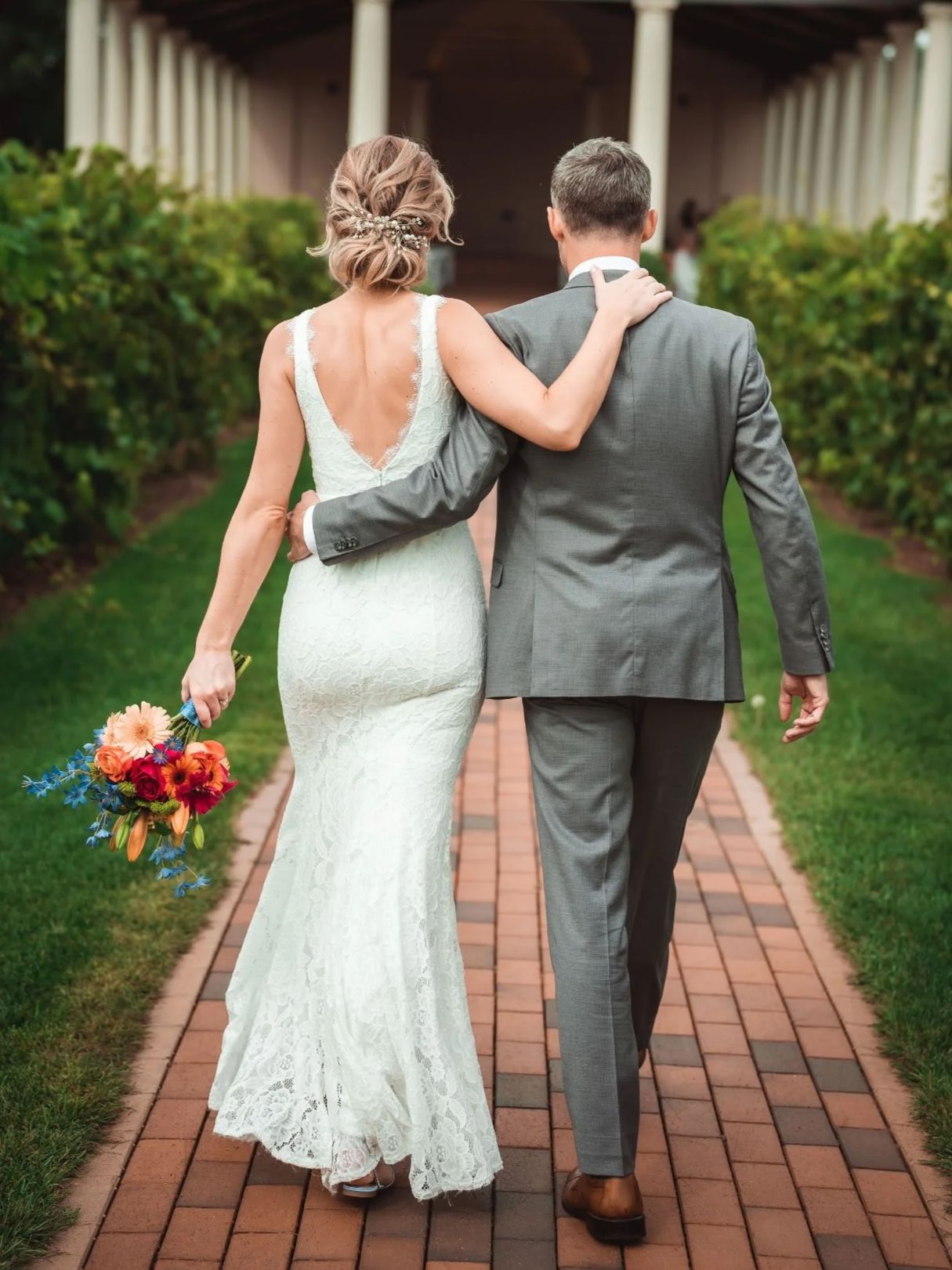 A bride and groom walking together on a brick path, with the bride's arm around the groom's shoulder. The bride is holding a colorful bouquet of flowers, and they are heading towards a building or pavilion.
