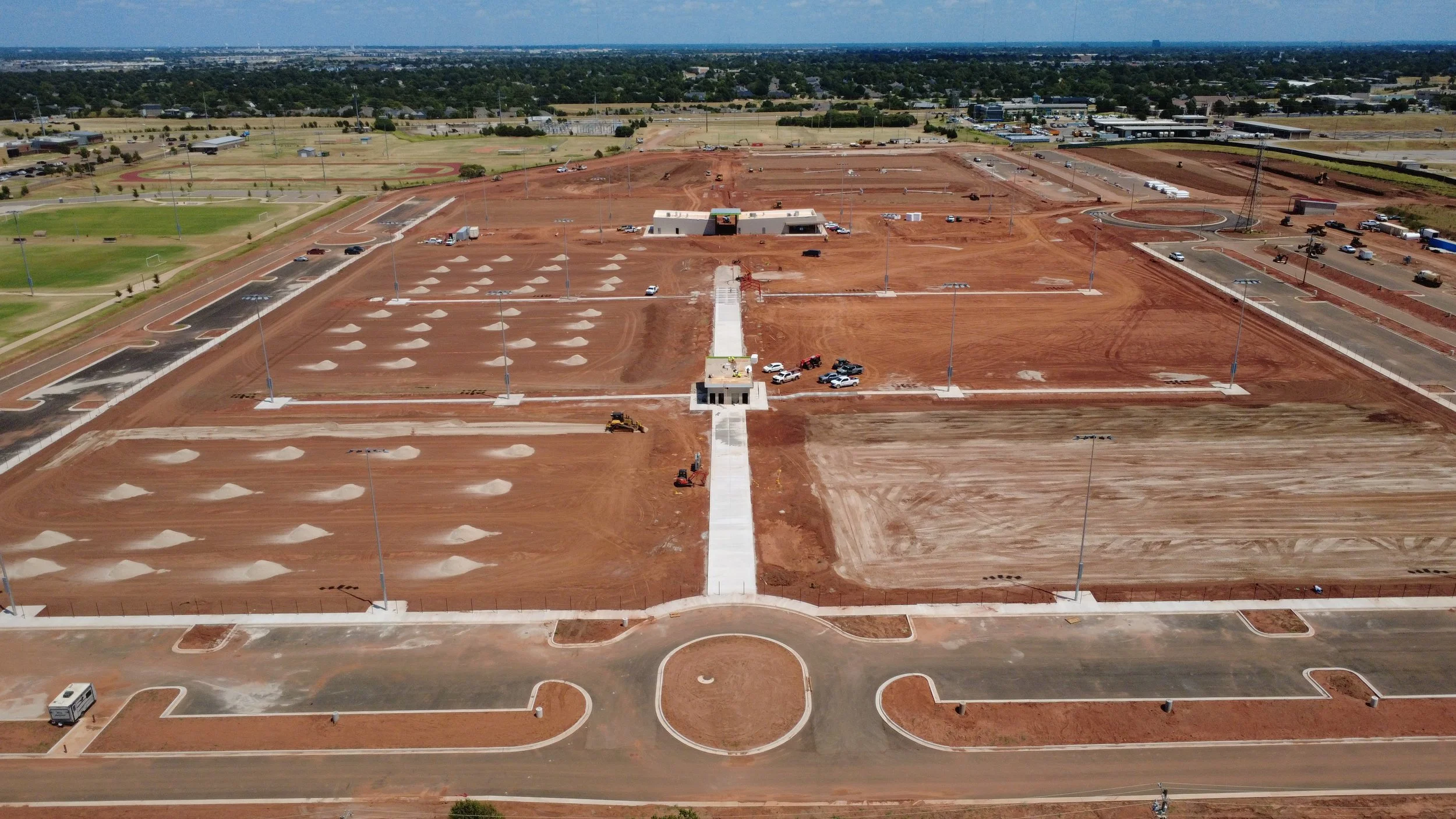 Aerial view of a construction site with earth-moving equipment and partially paved areas, surrounded by green fields and sports fields.