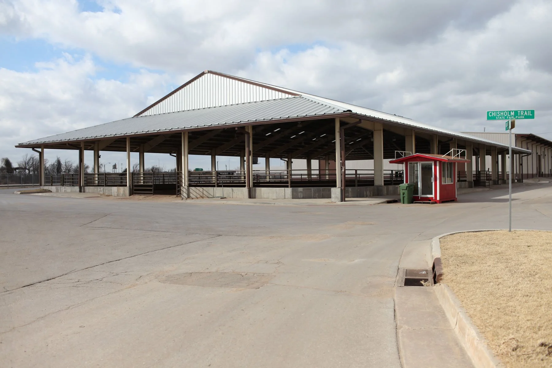 Empty horse riding arena with a metal roof, wooden railings, surrounded by paved areas and a grassy patch underneath a cloudy sky, with a sign reading 'Chilsholm Trail State Fair Park' and a red concession stand.