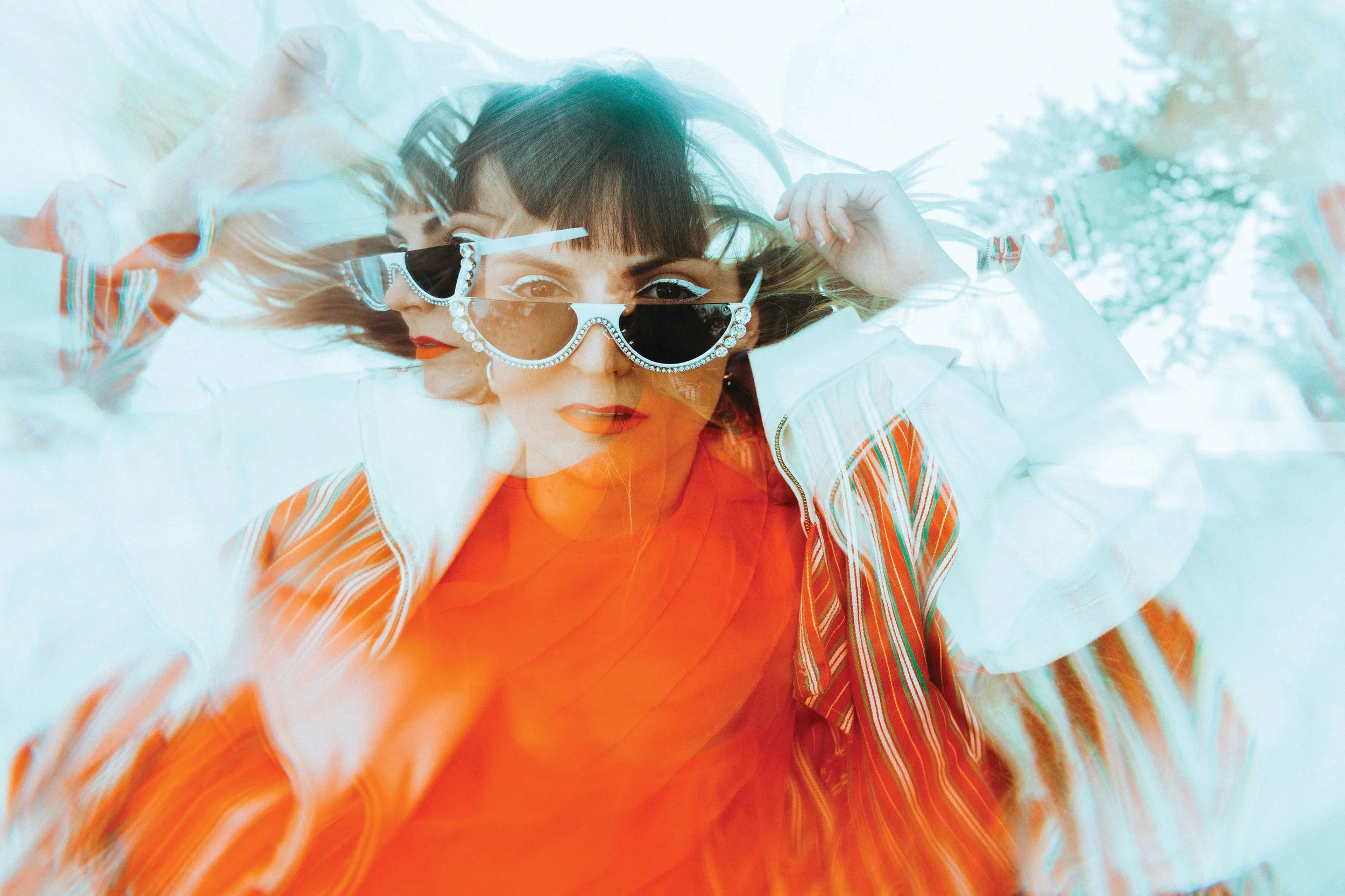 A woman with short dark hair and red lipstick wearing oversized sunglasses with rhinestone details, an orange shirt, and a white jacket with colorful stripes, holding her hair back with her hand, with a blurred outdoor background.