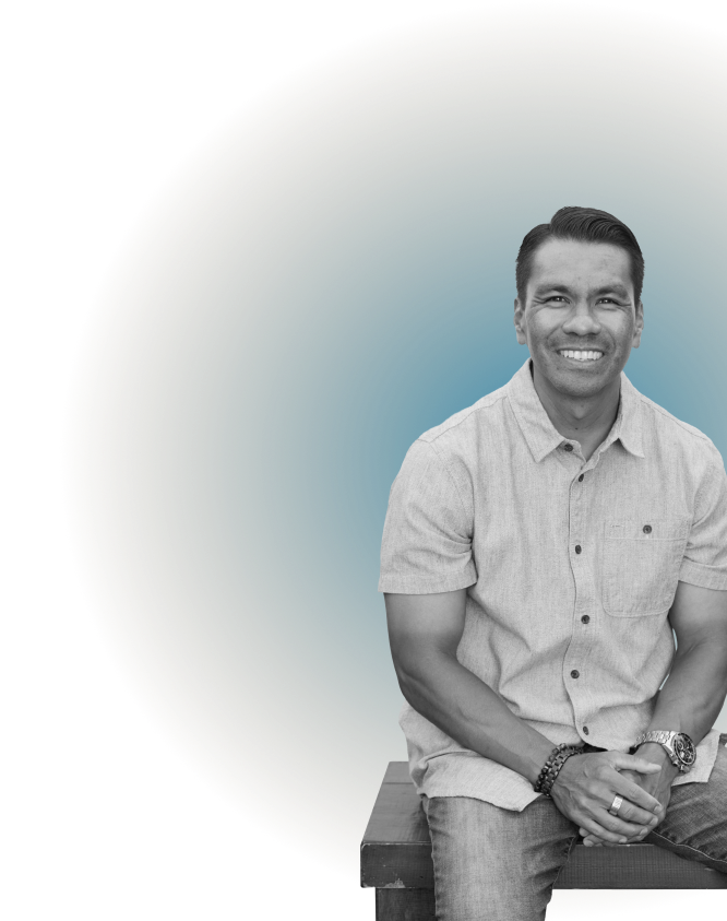 A smiling man in a short-sleeved button-up shirt, sitting on a wooden bench with his hands clasped, with a gradient colored background.