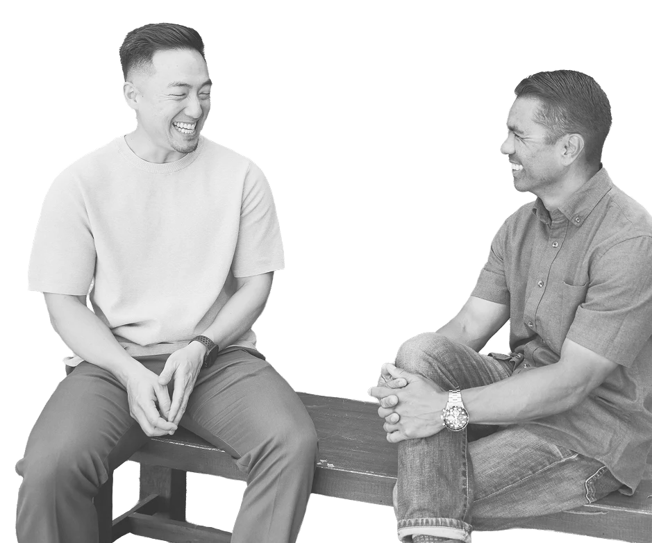 Two men sitting on a wooden bench, smiling and laughing at each other, engaging in a friendly conversation.