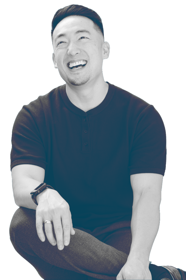 A man with short dark hair smiling, sitting with one arm resting on his knee, wearing a dark short-sleeved shirt and a watch on his wrist, set against a blue background.