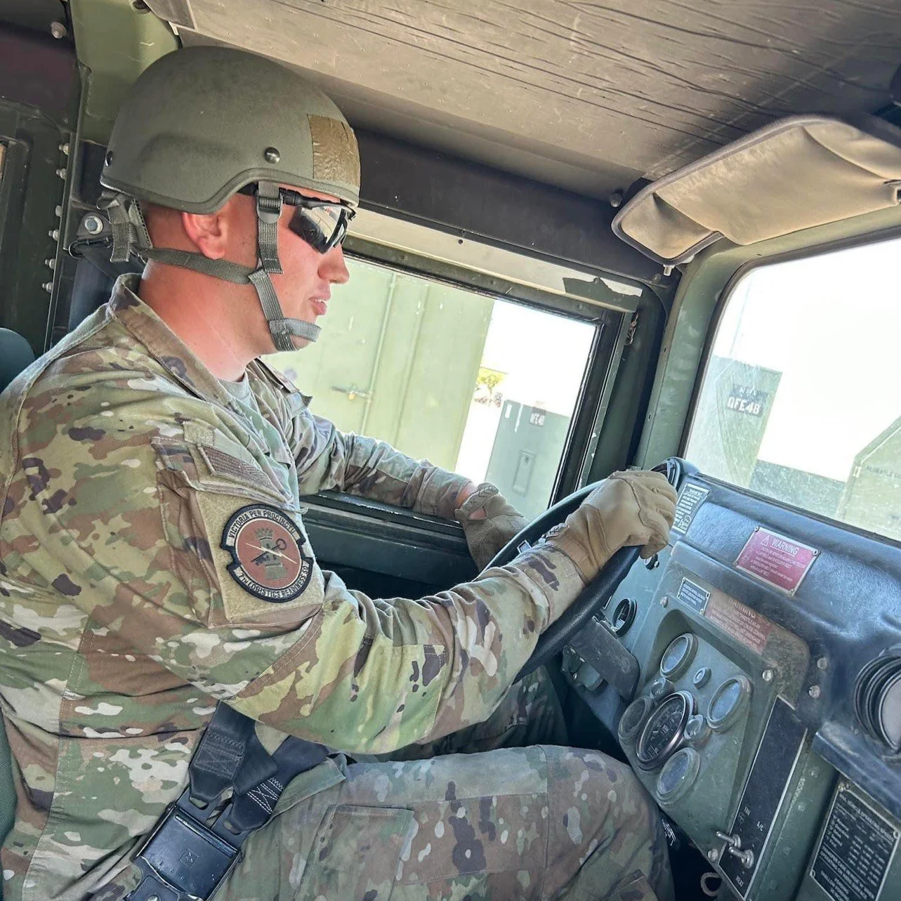 Badge and Rack Aligner, A airman in camouflage military uniform sitting in the driver’s seat of a military vehicle, wearing a helmet, sunglasses, and gloves.