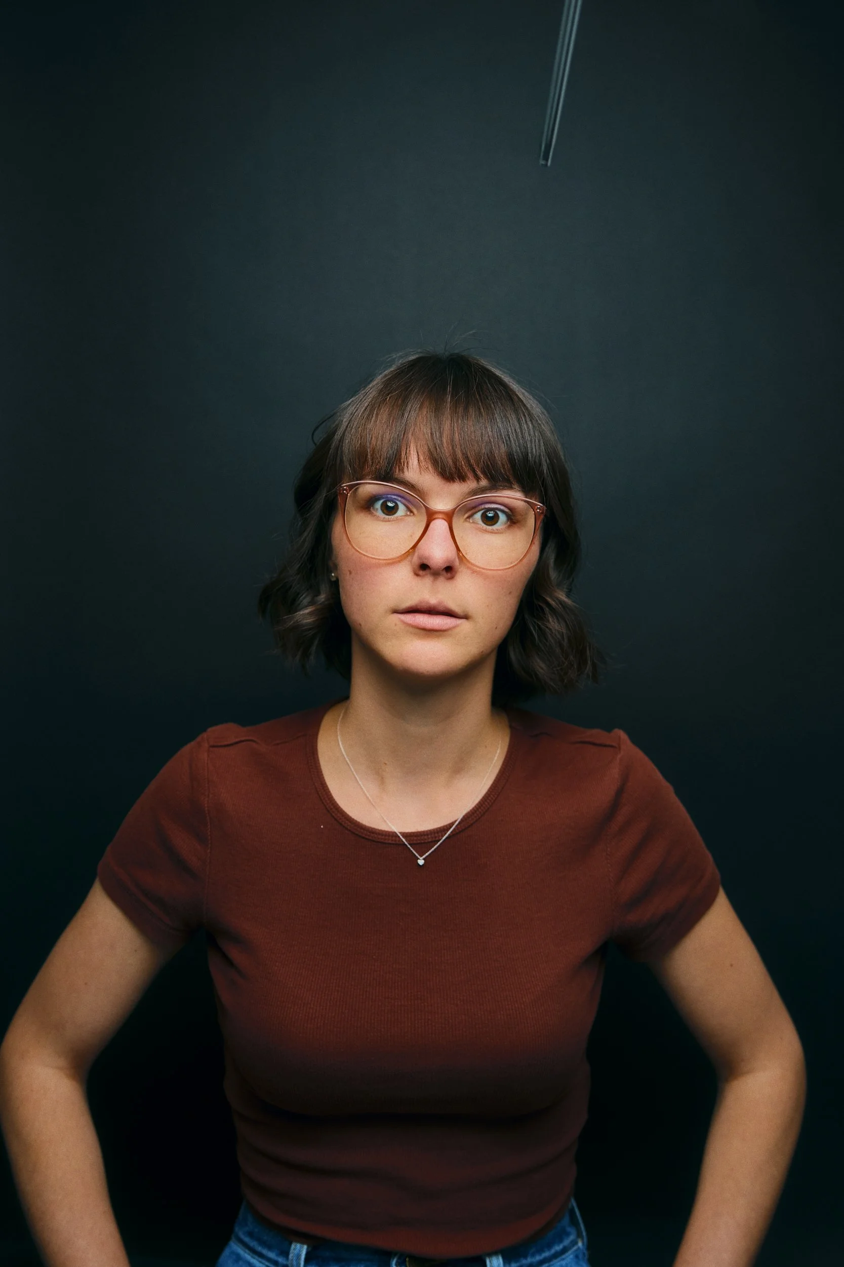 Junge Frau mit Brille, braunem T-Shirt und Halskette, vor dunklem Hintergrund, Blick direkt in die Kamera.