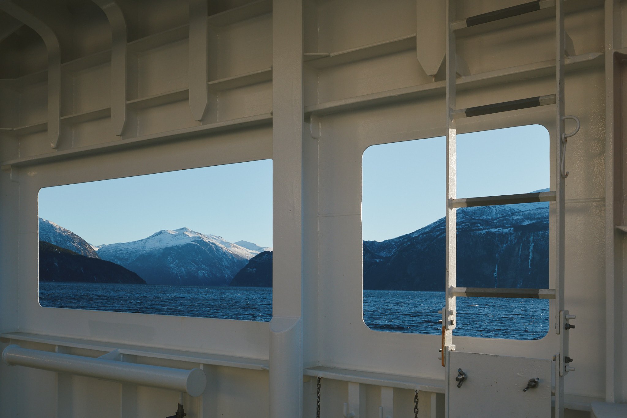 Blick durch eine Fensteröffnung eines Schiffs auf eine fjordartige Landschaft mit schneebedeckten Bergen unter einem klaren blauen Himmel.