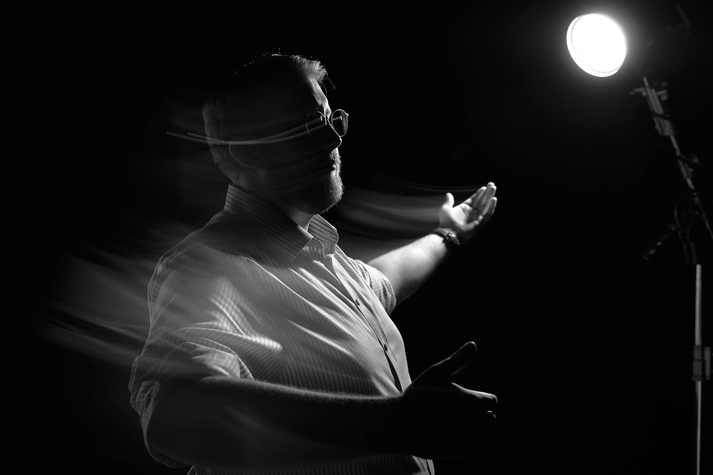 Ein Mann mit Bart und Brille steht vor einem Studio-Licht im schwarzen und weißen Bild. Er trägt ein Hemd und hat die Arme ausgebreitet.