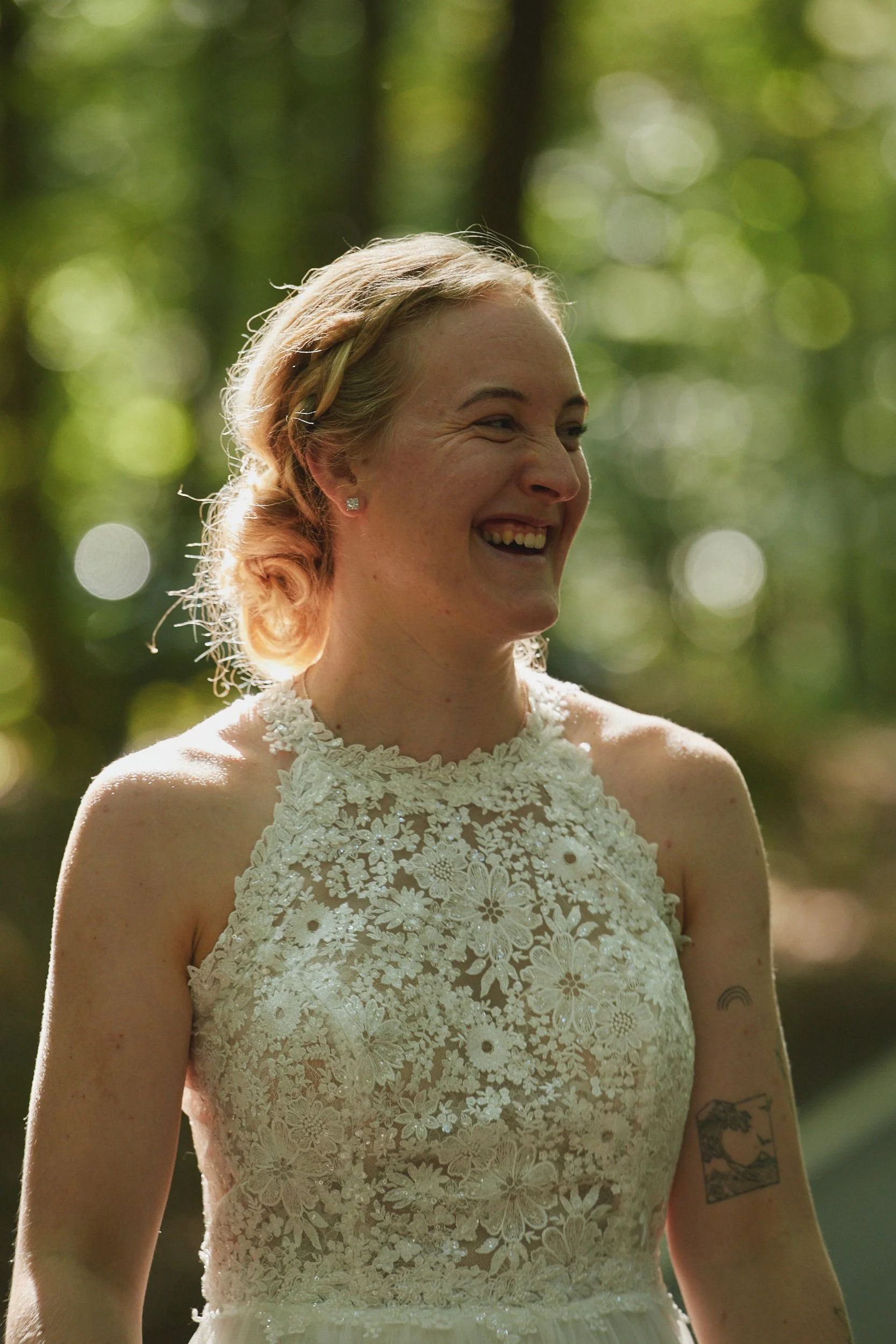 Eine lächelnde Frau trägt ein Brautkleid mit floralem Stickmuster und steht im Freien vor einem grünen, unscharfen Hintergrund.