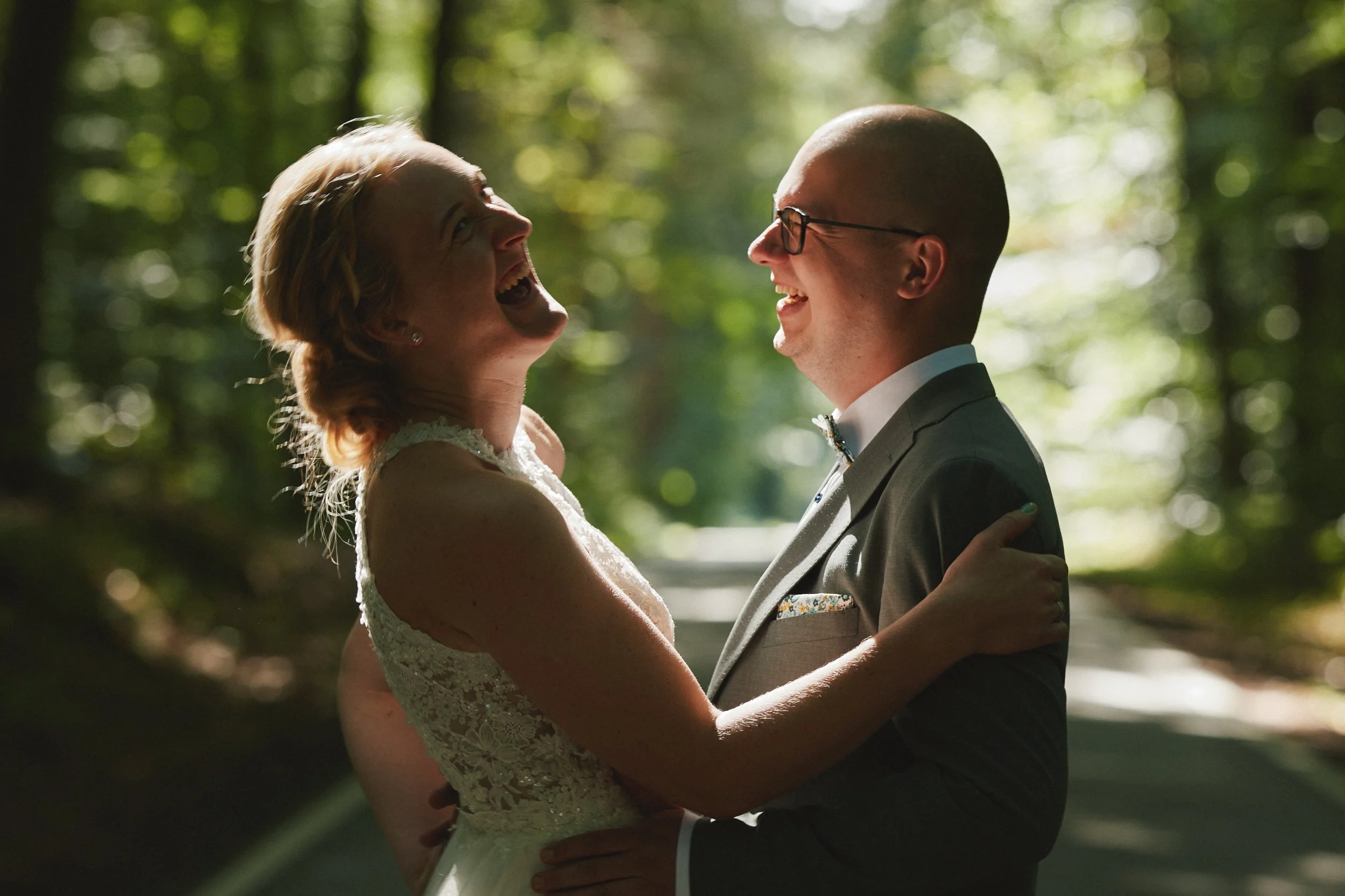 Ein Paar bei einer Hochzeit hält sich im Sonnenlicht im Wald und lacht glücklich.