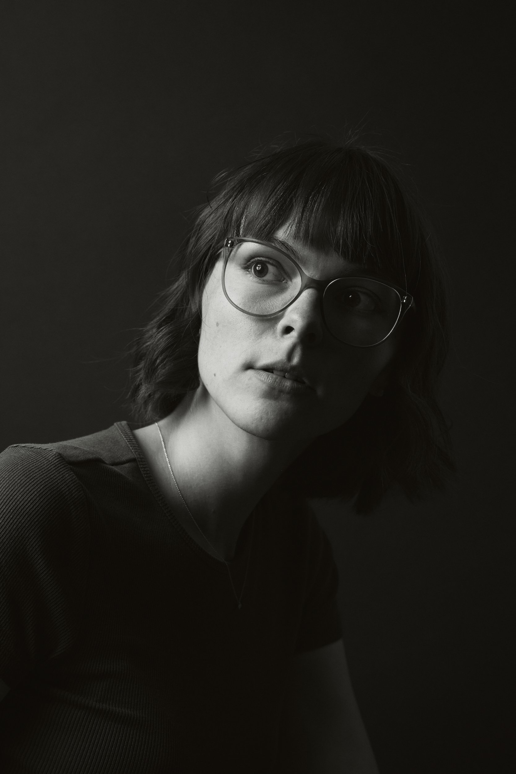 Schwarz-Weiß-Porträt einer jungen Frau mit Kurzhaarfrisur, Brille und einem grauen T-Shirt, Blick nach links, dunkler Hintergrund.