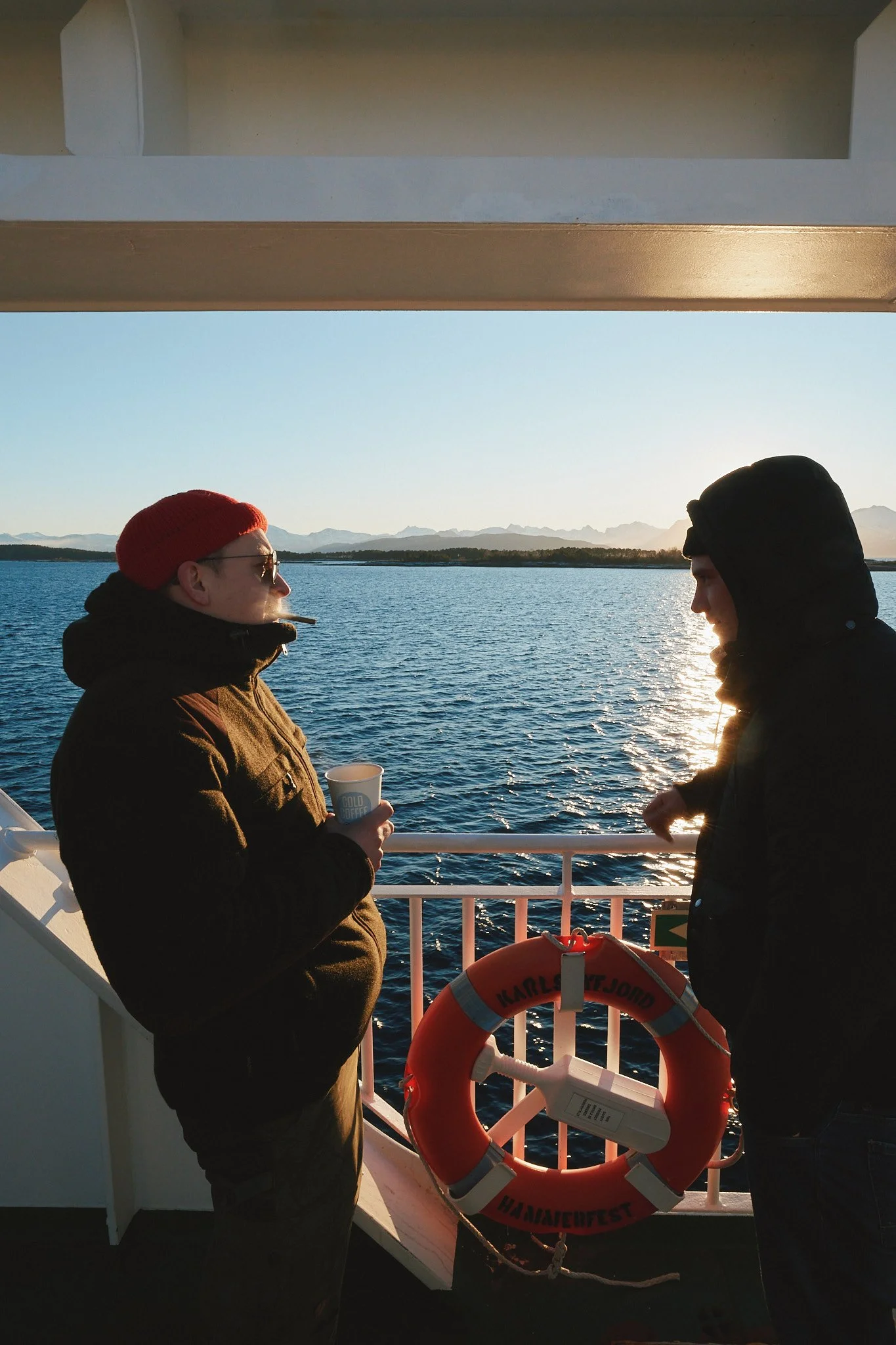 Zwei Personen stehen auf dem Deck eines Schiffes mit Blick auf das Wasser und Berge im Hintergrund. Eine Person trägt eine rote Mütze und hält eine Tasse, die andere trägt eine schwarze Kapuze. Es ist Sonnenuntergang.