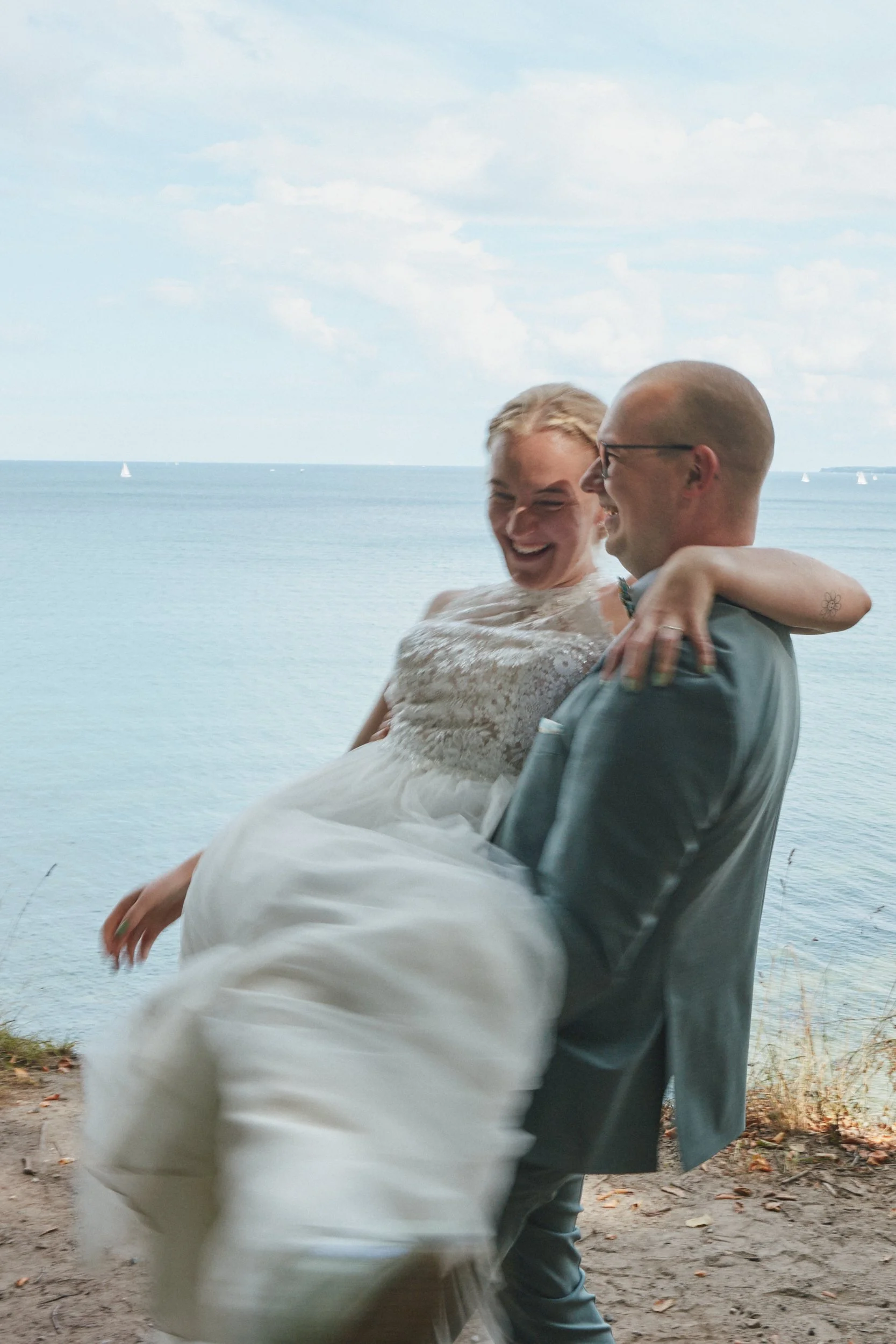 Ein Mann und eine Frau, die sich freuen, bei einem Treffen am Wasser. Die Frau trägt ein weißes Kleid und der Mann trägt einen grauen Anzug. Sie umarmen sich lachend und glücklich.