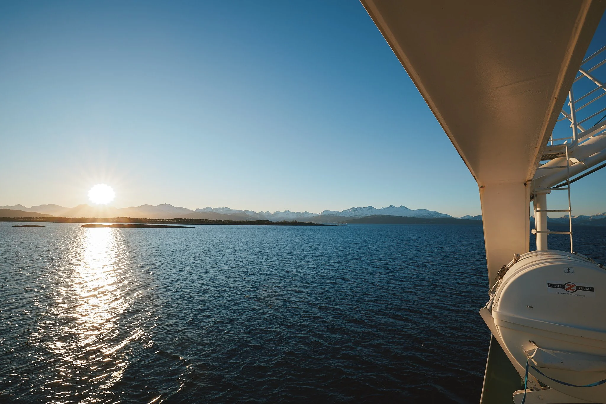Blick vom Deck eines Schiffes auf einen See, mit Bergen im Hintergrund und der Sonne die kurz vor dem Horizont untergeht. Im rechten Bildteil ist ein Teil des Schiffsdachs sichtbar.