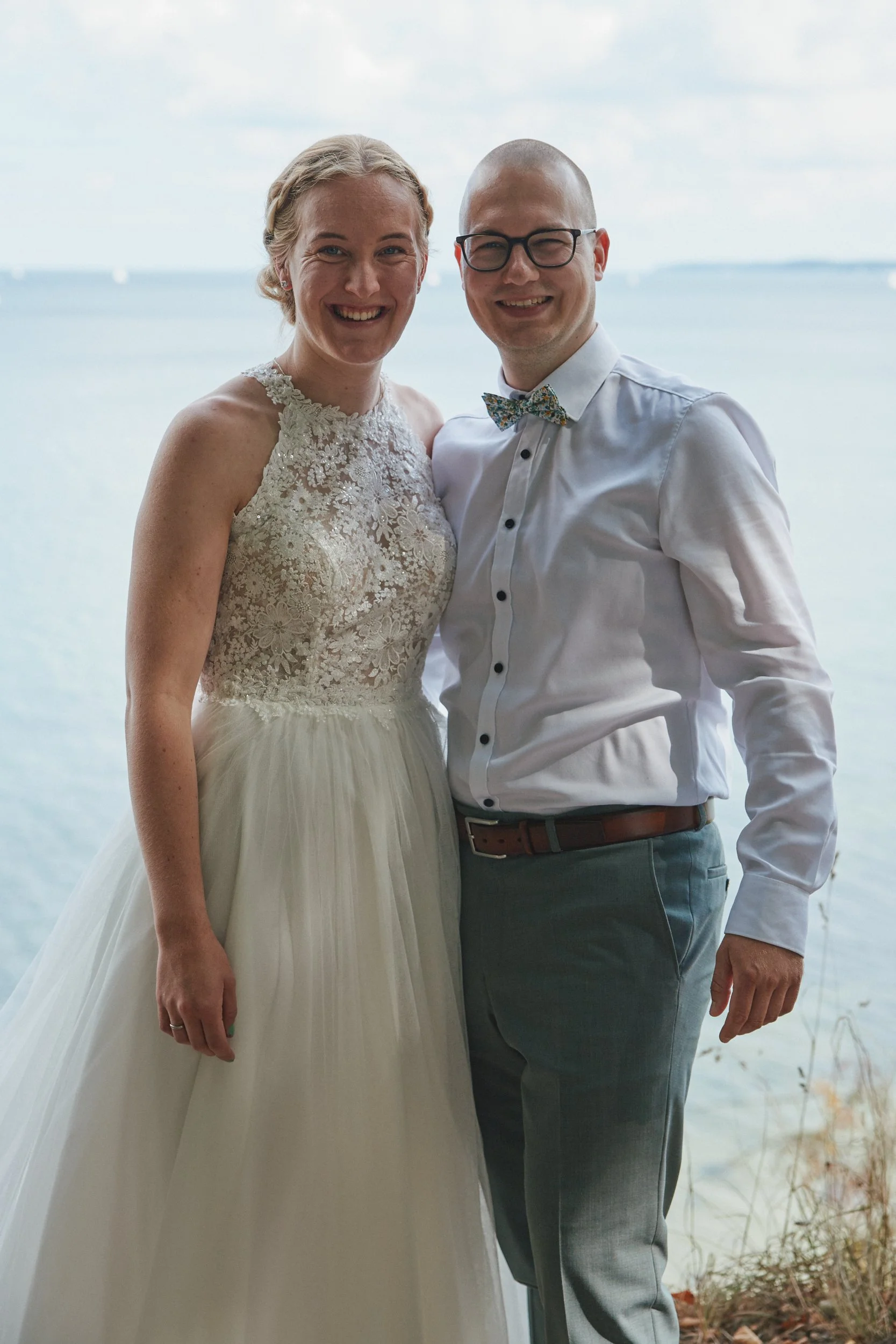 Ein lachendes Brautpaar steht vor einem See, die Frau trägt ein weißes Hochzeitskleid mit Spitze, der Mann ein weißes Hemd mit Fliege.