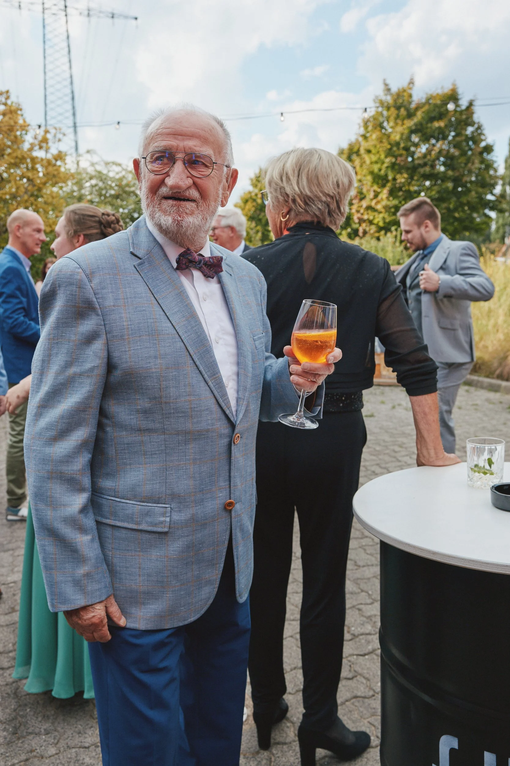 Älterer Mann in einem bläulichen Anzug mit Fliege hält ein Weinglas bei einer Gartenparty. Im Hintergrund stehen Menschen, einige in Anzügen, im Gespräch, umgeben von Bäumen und einem blauen Himmel.