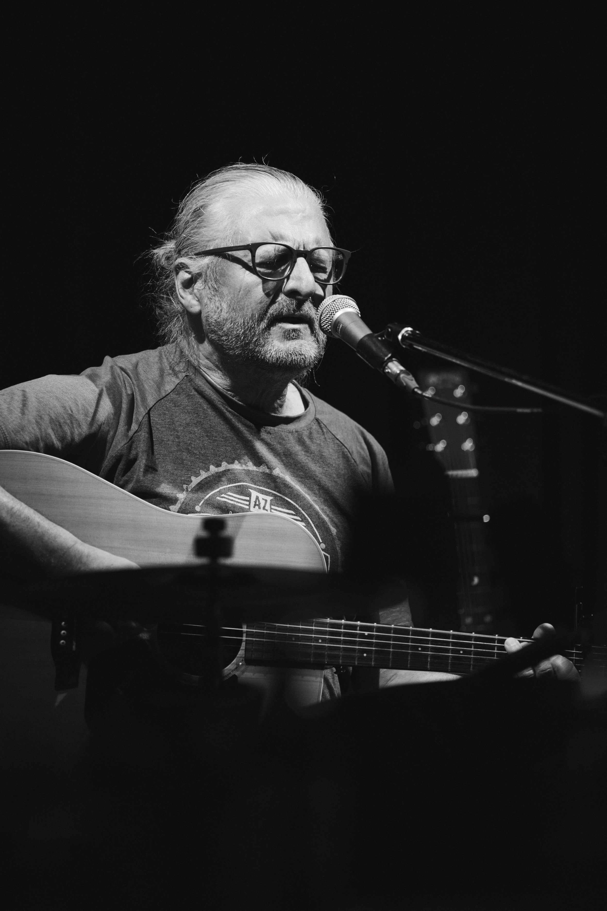 Schwarz-weiß-Foto eines Mannes, der Gitarre spielt und vor einem Mikrofon singt, während er eine Brille trägt.