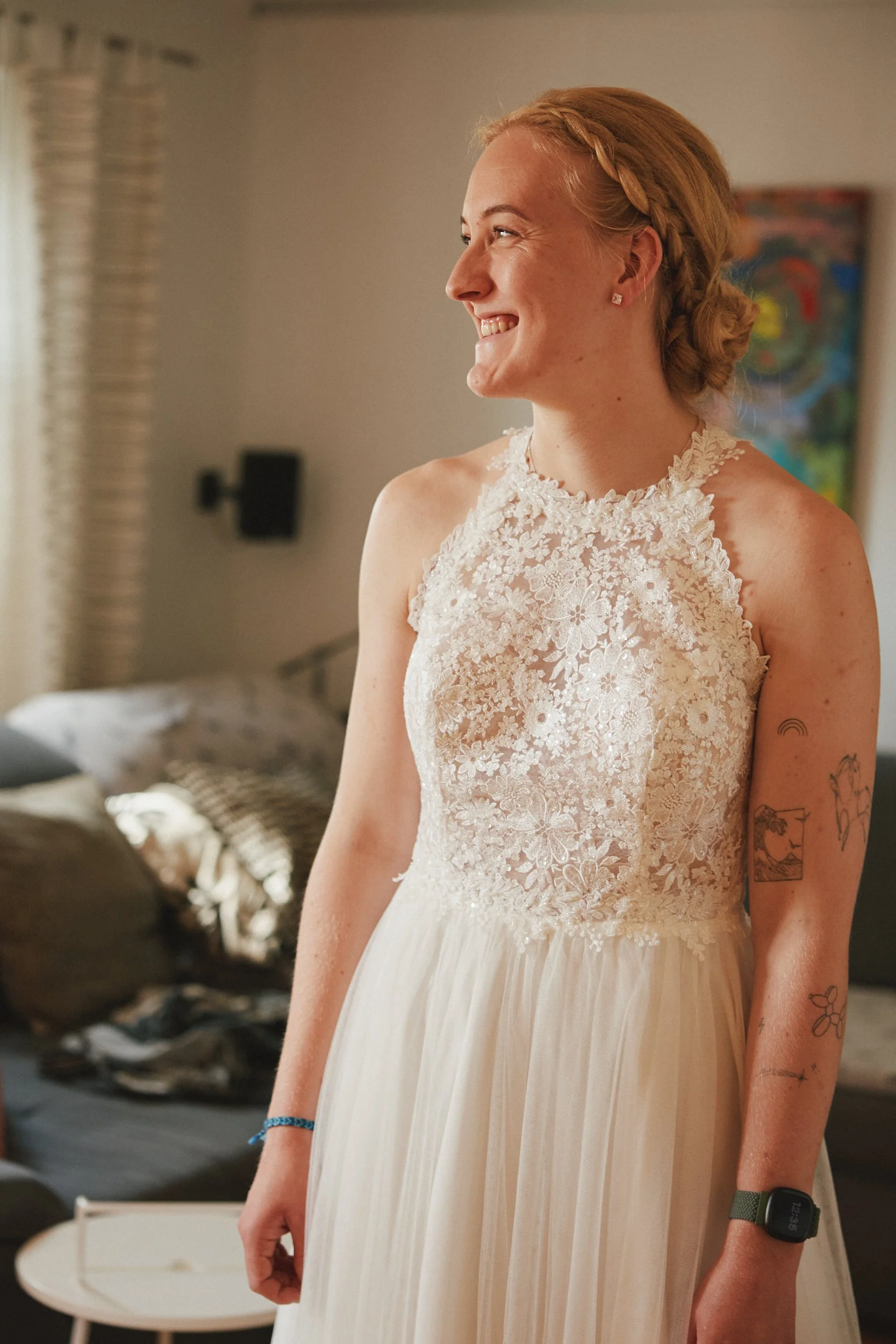 Junge Frau in weißem Brautkleid lächelnd, Steh in einem Wohnzimmer, Tattoos am Arm, trägt eine Armbanduhr und einen blauen Armband, im Hintergrund ein Sofa und ein erschaffenes Gemälde