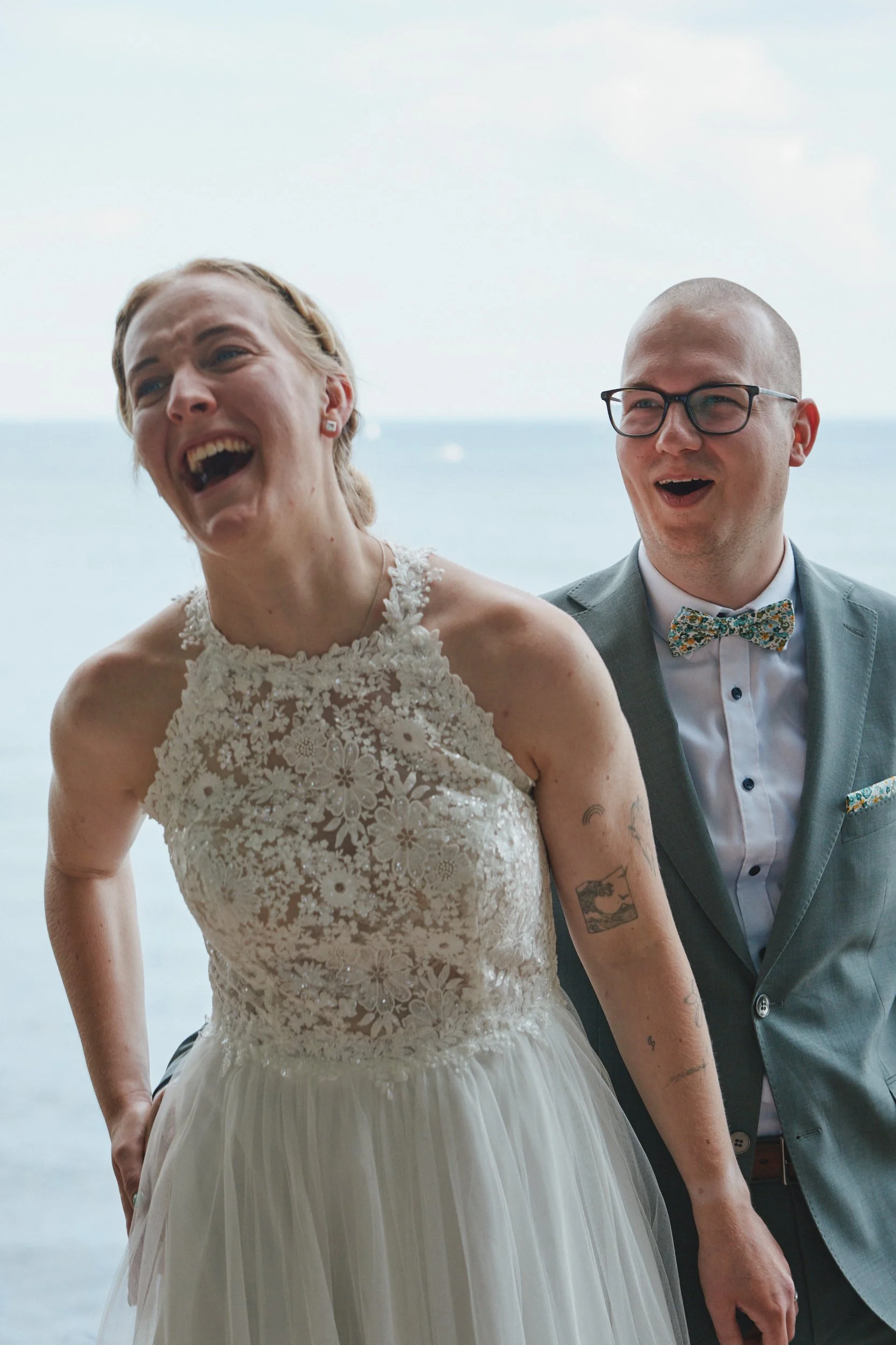 Ein lachendes Paar bei einer Hochzeit am Strand, die Frau trägt ein weißes Spitzenkleid und hat Tattoos am Arm, der Mann trägt eine Brille und einen grauen Anzug mit Fliege.