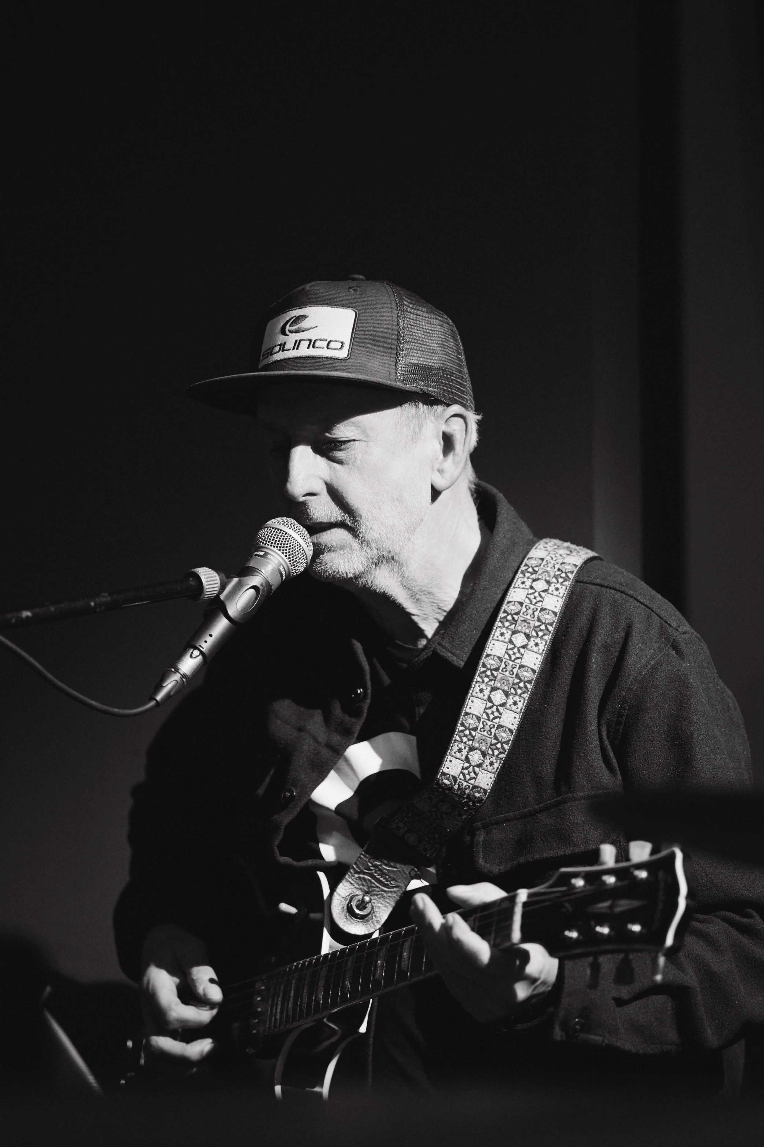 Ein älterer Mann mit grauem Bart spielt Gitarre und singt in ein Mikrofon, trägt eine Cap mit dem Logo 'SOLINCO' und eine Jacke, im dunklen Hintergrund.