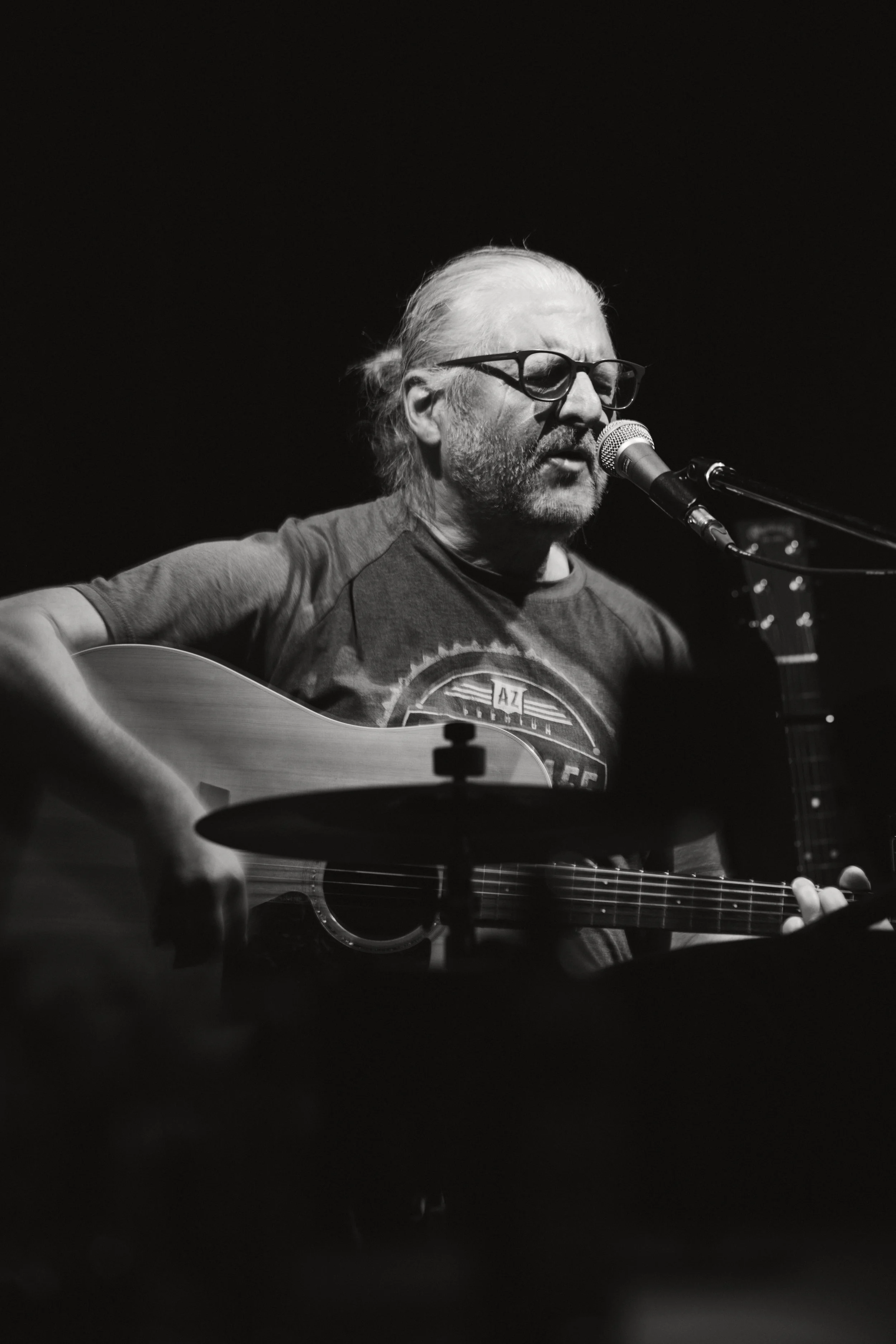 Ein älterer Mann mit Brille singt in Mikrofon und spielt Gitarre auf einer Bühne, schwarz-weiß.