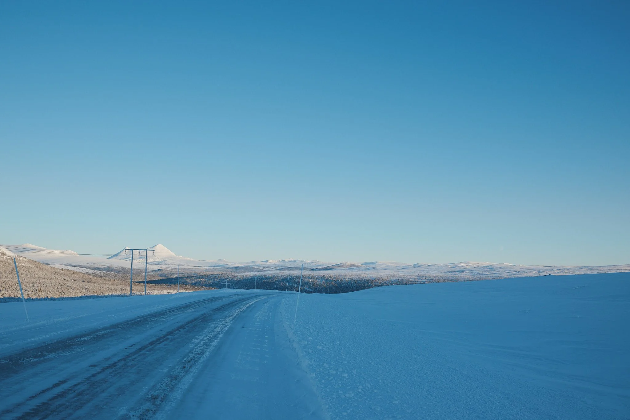 Eine verschneite Landschaft mit klarer blauer Himmel, eine Straße ist sichtbar mit Schneefeldern auf beiden Seiten, und im Hintergrund erkennen man Berge.