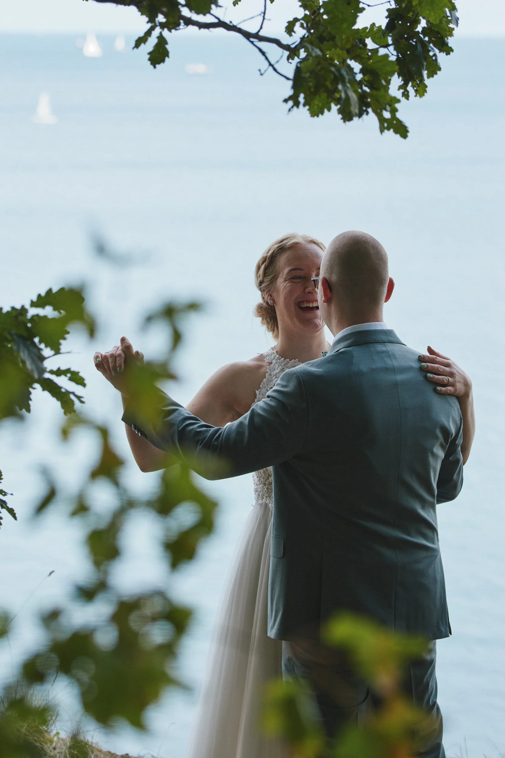 Ein Brautpaar tanzt am Wasser, umgeben von Bäumen, beim Blick durch Blätter, bei einer Hochzeit im Freien.