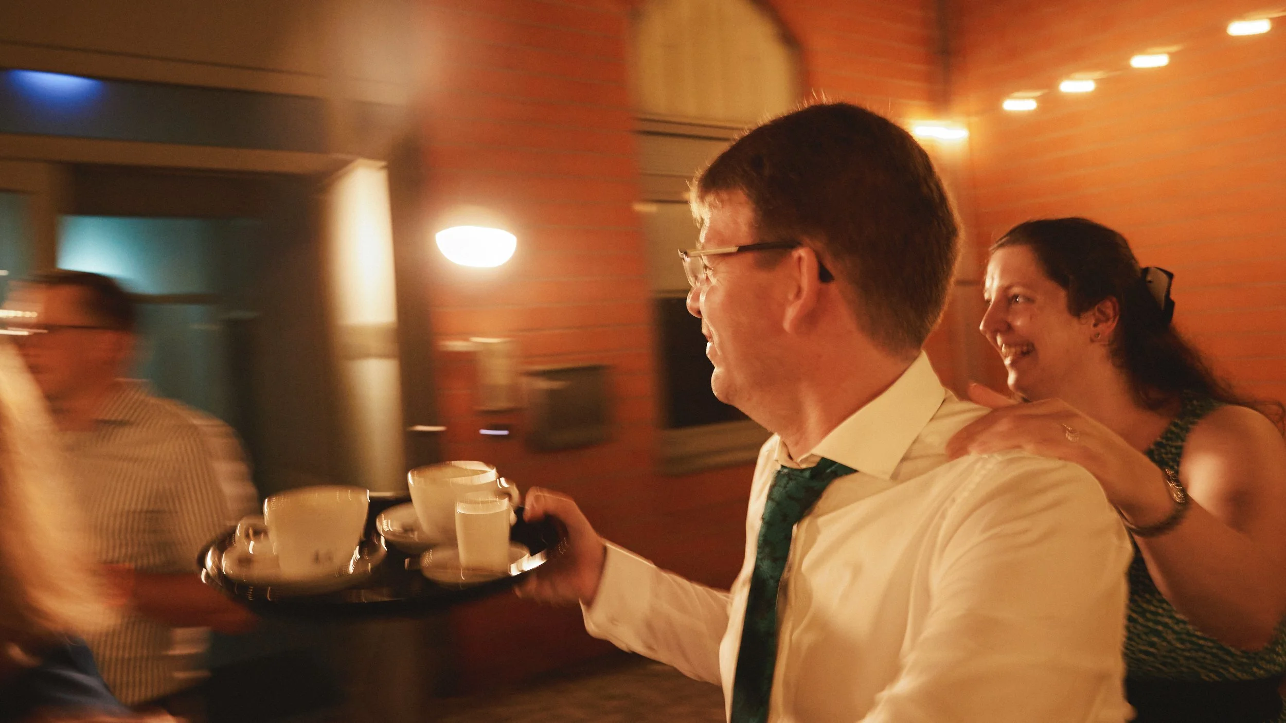 Ein Mann mit Brille und weißem Hemd hält mehrere Kaffeetassen auf einem Tablett, während er in einem festlichen Raum mit roten Ziegelwänden ist. Es ist eine fröhliche Atmosphäre mit anderen Leuten im Hintergrund.