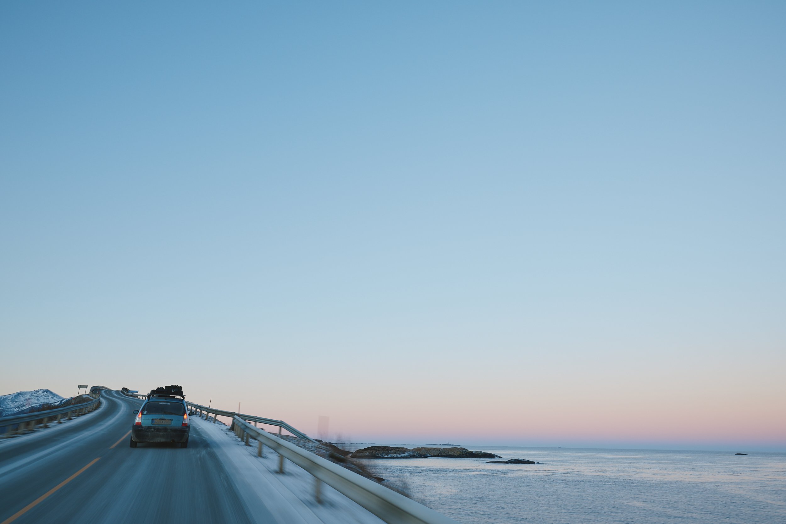Ein Auto fährt auf einer kurvigen Straße entlang der Küste bei Sonnenuntergang, mit Blick auf das Wasser und den Himmel.