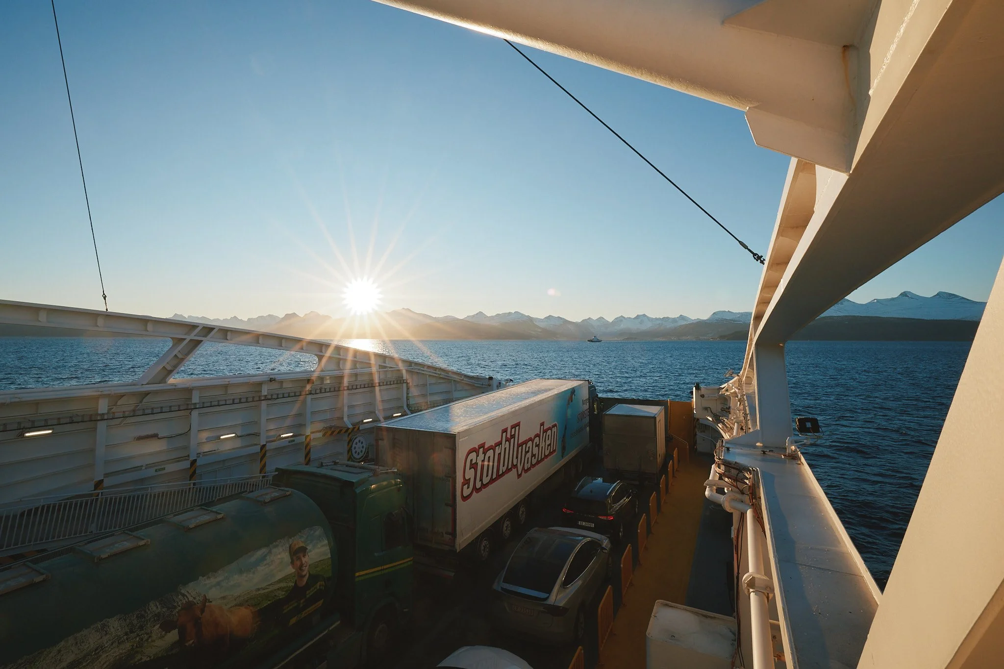 Blick von einem Schiff auf das Meer mit schneebedeckten Bergen im Hintergrund, die Sonne steht niedrig am Himmel.