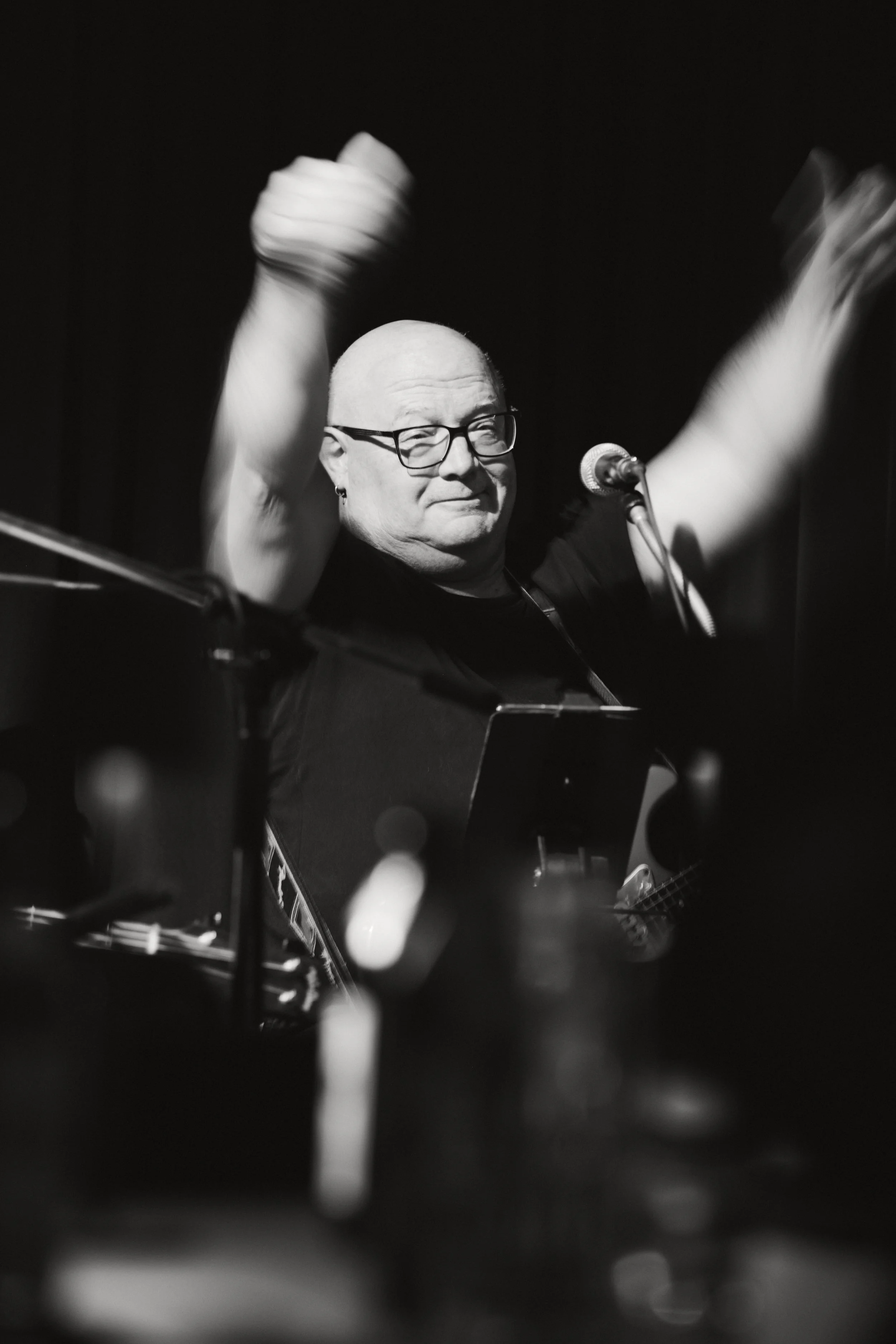 Schwarz-weiß-Foto eines Mannes mit Brille, der ein Musikinstrument spielt, vor einem Mikrofon, in einer dunklen Umgebung, möglicherweise bei einem Auftritt oder in einem Studio.
