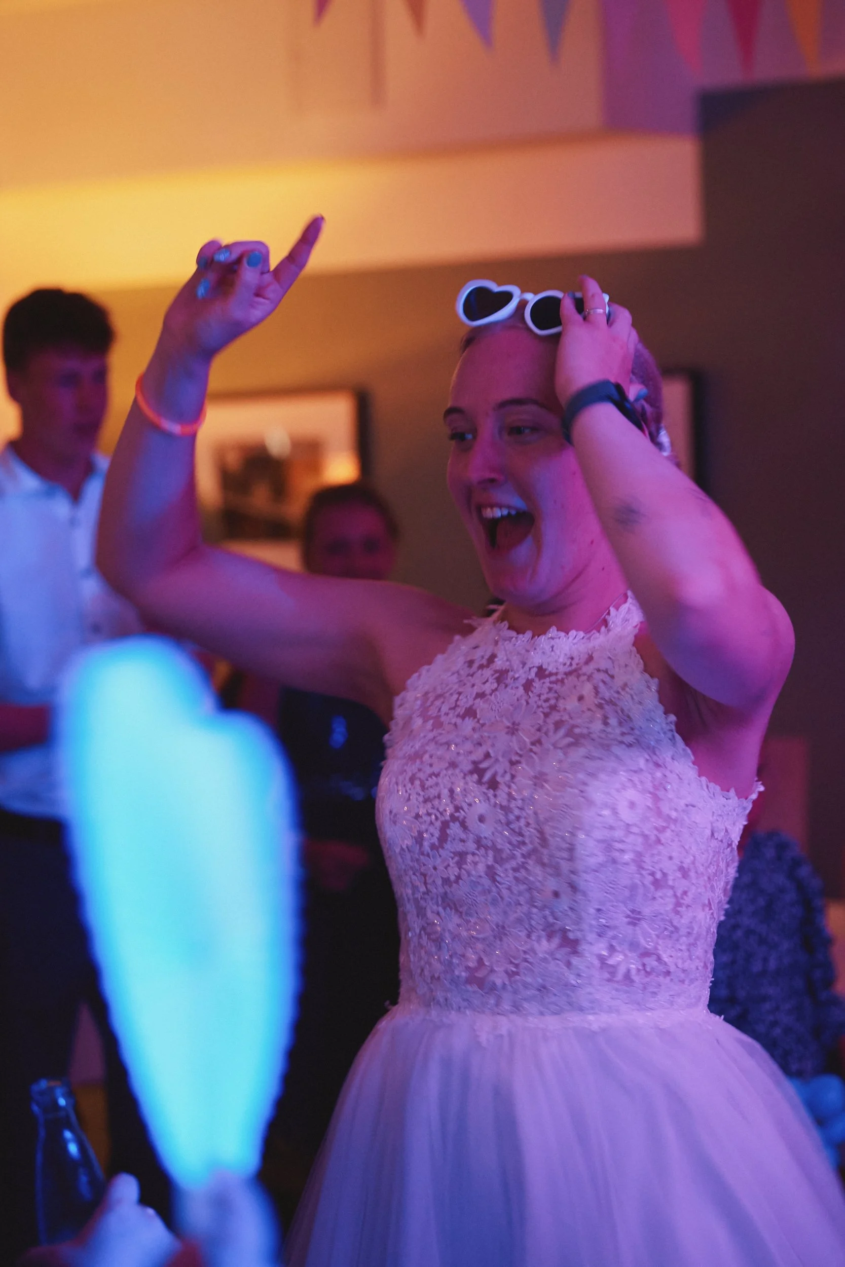 Junge Frau mit Herz-Linsenbrille, tanzt bei einer Party in einem Raum mit bunten Lichter. Im Hintergrund sind andere Menschen zu sehen.