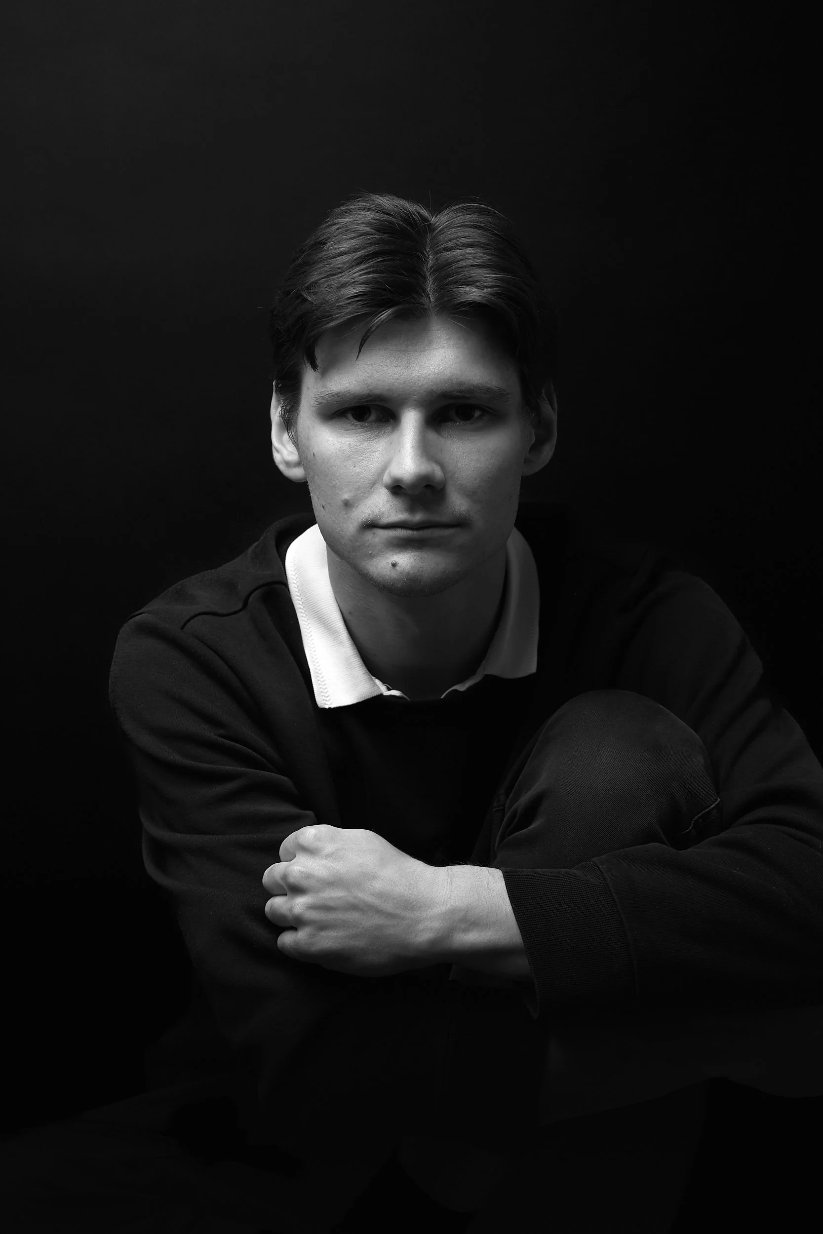 Schwarz-weiß-Foto eines jungen Mannes, der nach vorne schaut, mit dunklem Hintergrund. Er trägt eine dunkle Jacke und ein weißes Hemd.