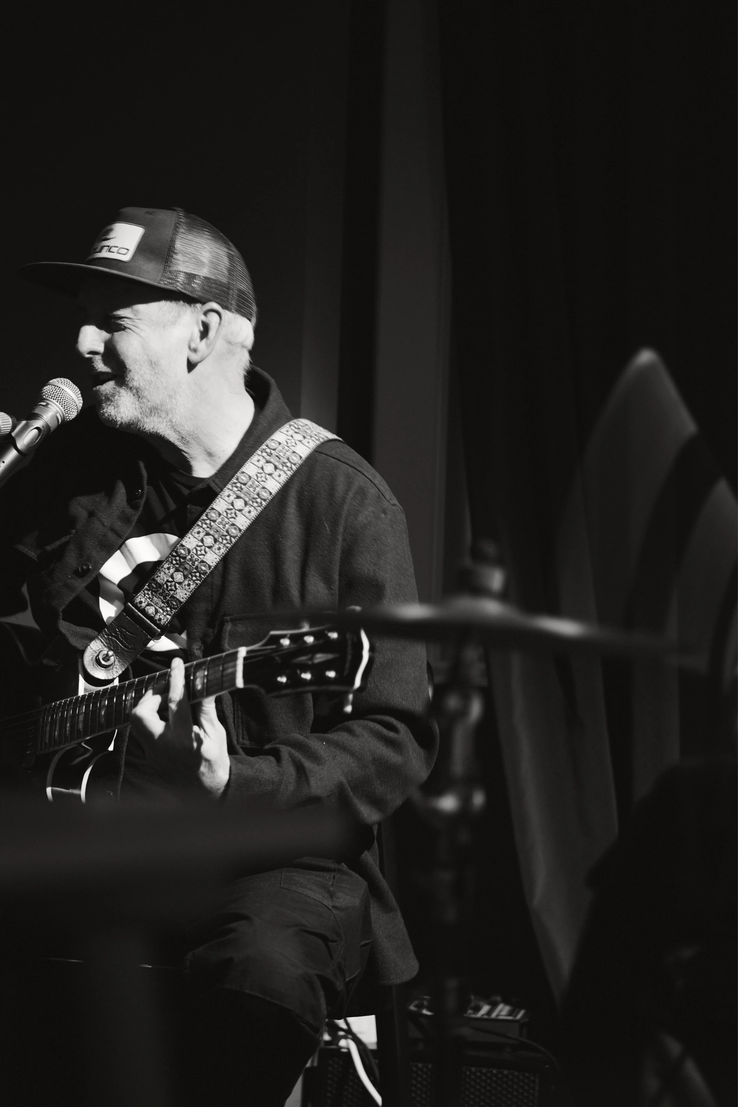 Ein Mann spielt Gitarre und singt in einem dunklen Raum, trägt einen Baseballcap und eine Jacke.