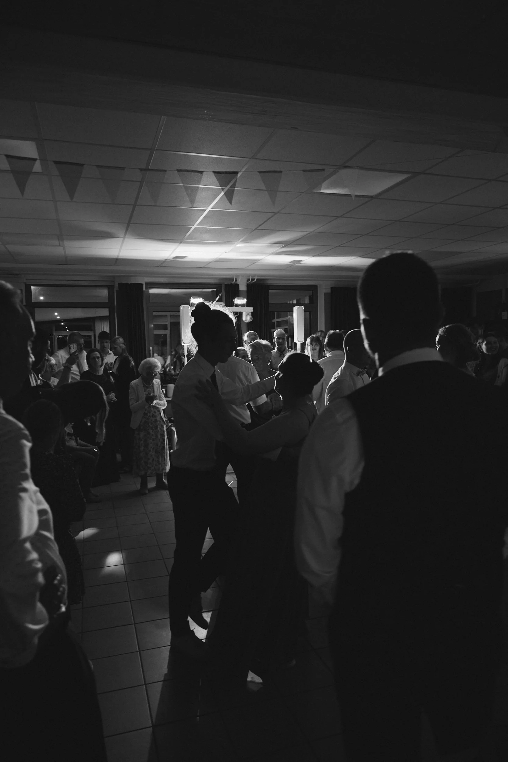 Schwarz-Weiß-Foto einer Hochzeit mit mehreren Personen, die umeinander tanzen, im Raum mit Deckenlampen und Fenstern im Hintergrund.