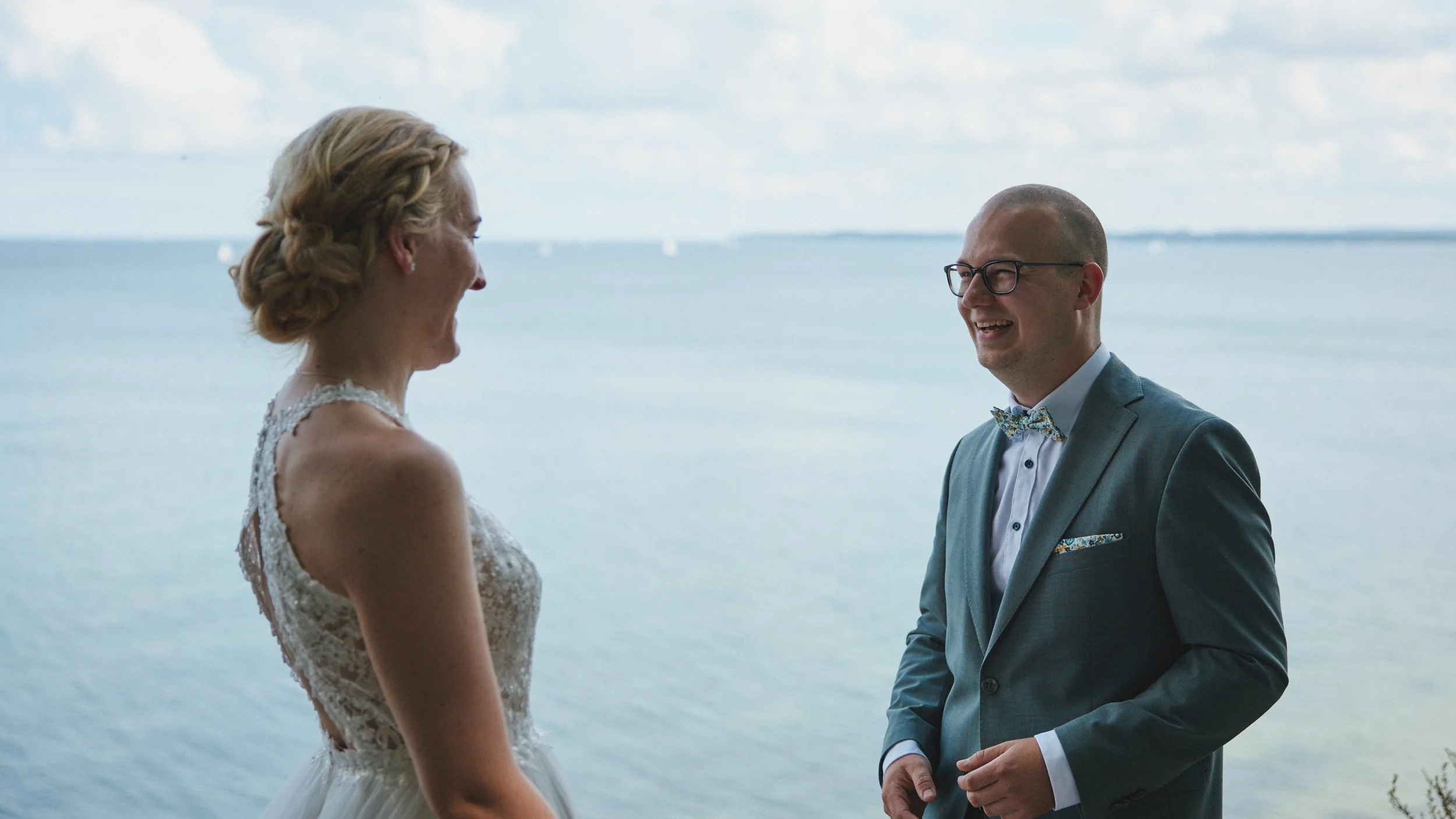 Ein Brautpaar steht am Wasser, die Frau trägt ein Hochzeitskleid mit Spitze, der Mann trägt einen grauen Anzug mit Fliege. Sie lachen sich an, im Hintergrund ist das Meer mit kleinen Segelbooten und Himmel mit Wolken zu sehen.