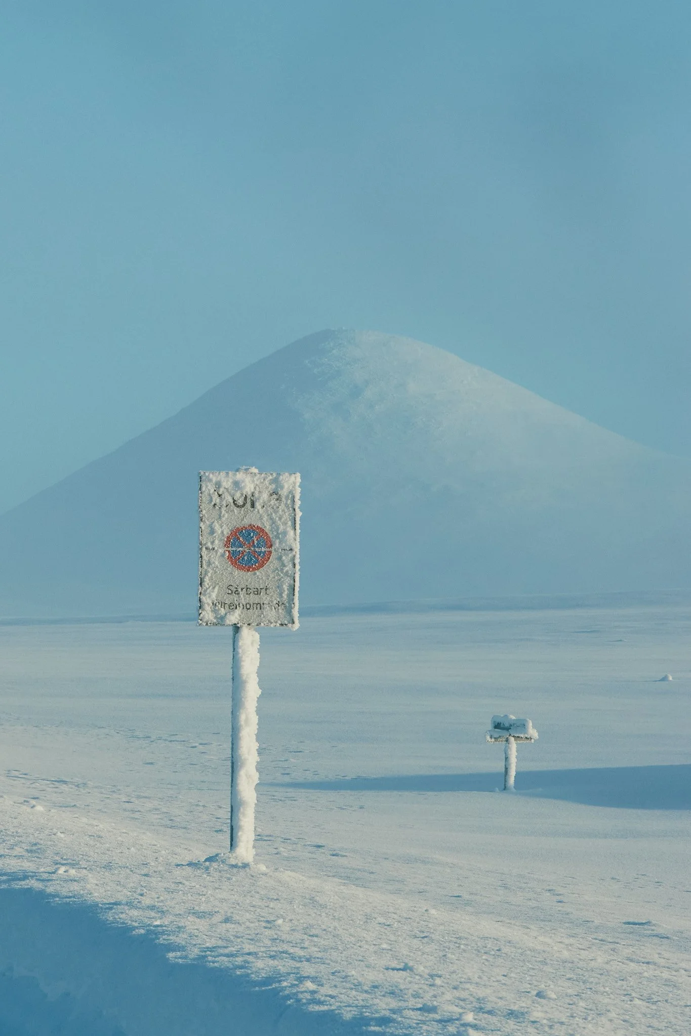 Schneebedeckter Zustand auf einer offenen Fläche mit einem Wegweiser und einem weiteren Schild im Schnee, im Hintergrund schneeweißer Berg unter klarem blauen Himmel.