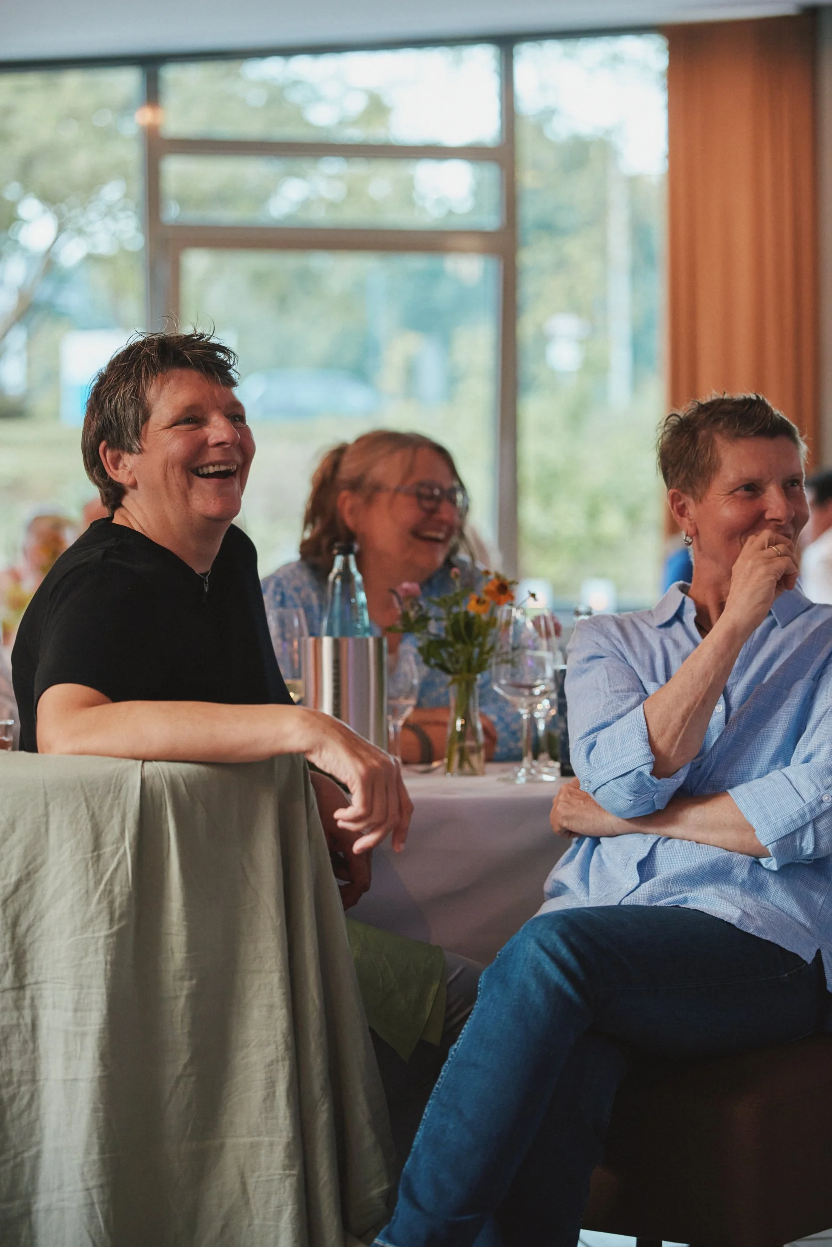 Menschen lachen und unterhalten sich bei einem Treffen in einem Raum mit großen Fenstern und natürlichem Licht.
