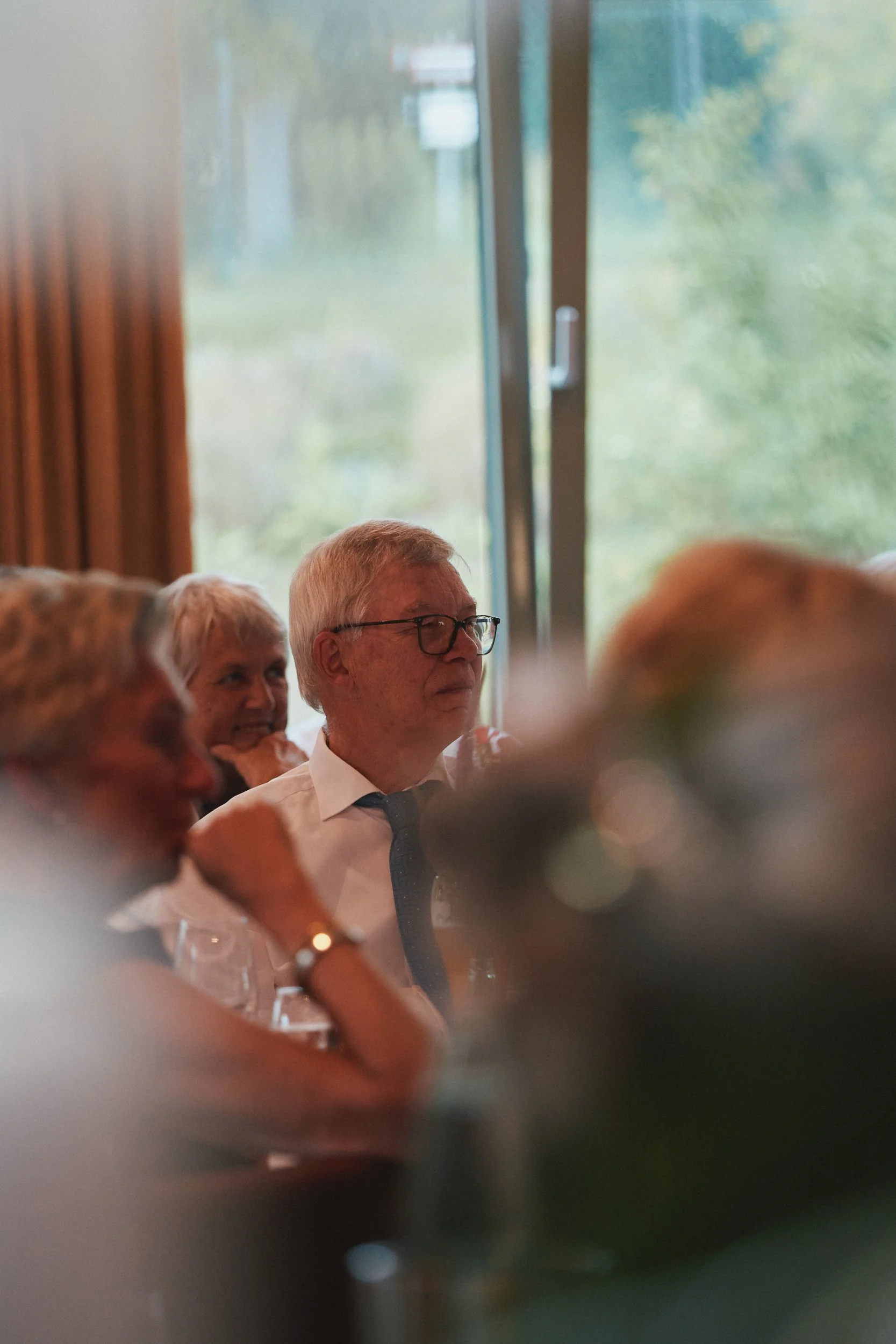 Ein Mann mit Brille in einem weißen Hemd und Krawatte sitzt bei einem Treffen mit anderen Personen in einem Raum mit großen Fenstern und grüner Umgebung im Hintergrund.