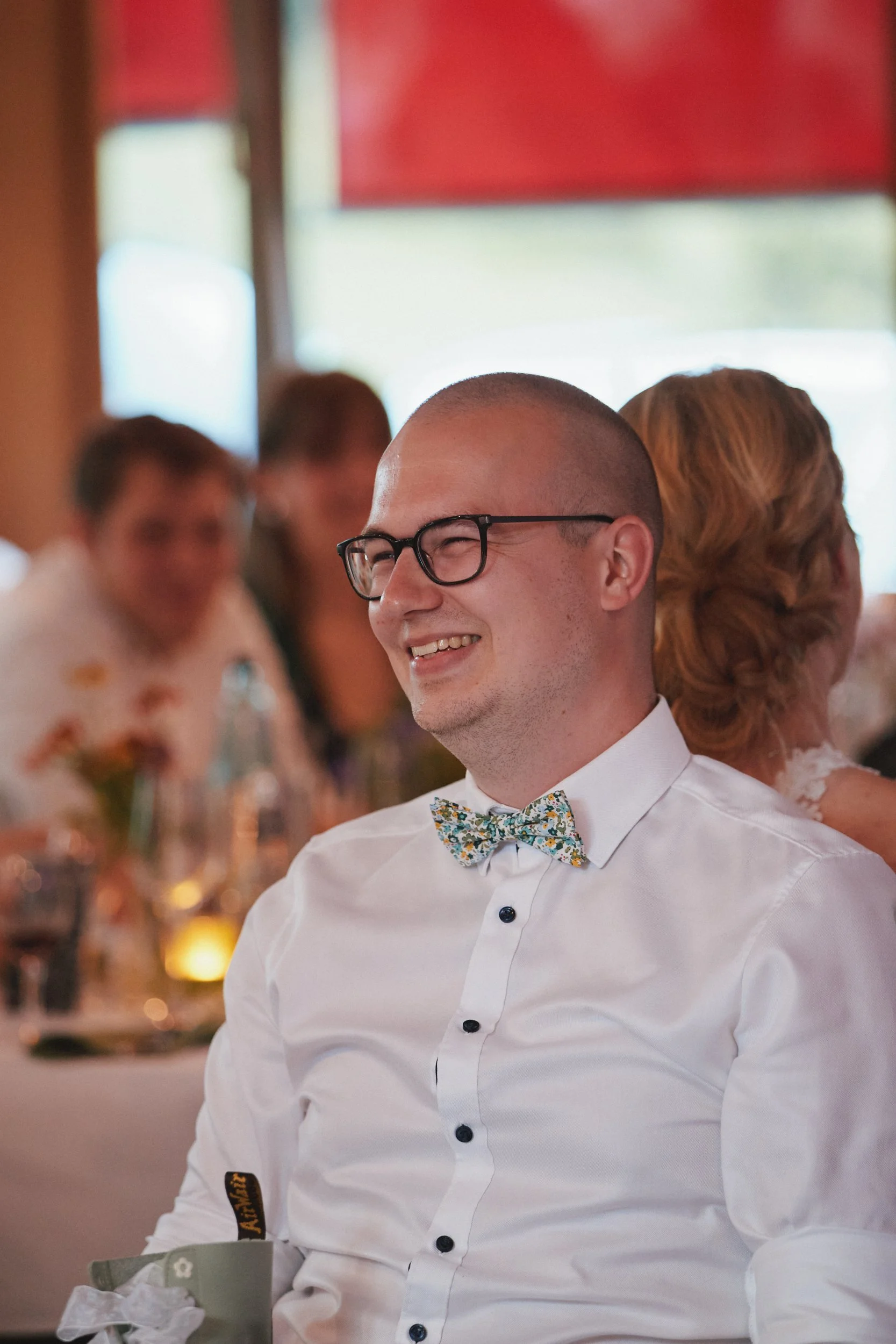 Junge Mann mit Brille, weißem Hemd und buntem Fliege, lächelnd in einem festlichen Anzug, im Hintergrund Gäste während einer Feier in einem Restaurant oder Saal.