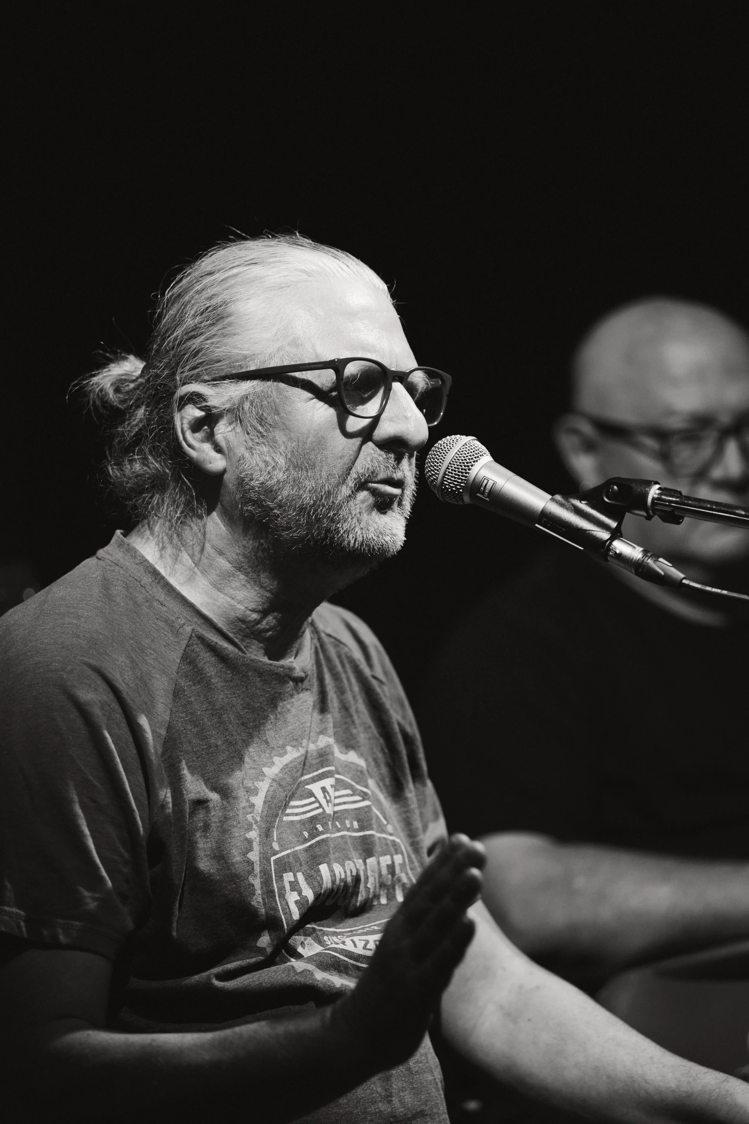 Schwarz-weiß Foto eines älteren Mannes mit weißen Haaren, Brille und grauem Bart, der singt oder spricht vor einem Mikrofon auf einer Bühne, mit einem weiteren Mann im Hintergrund.