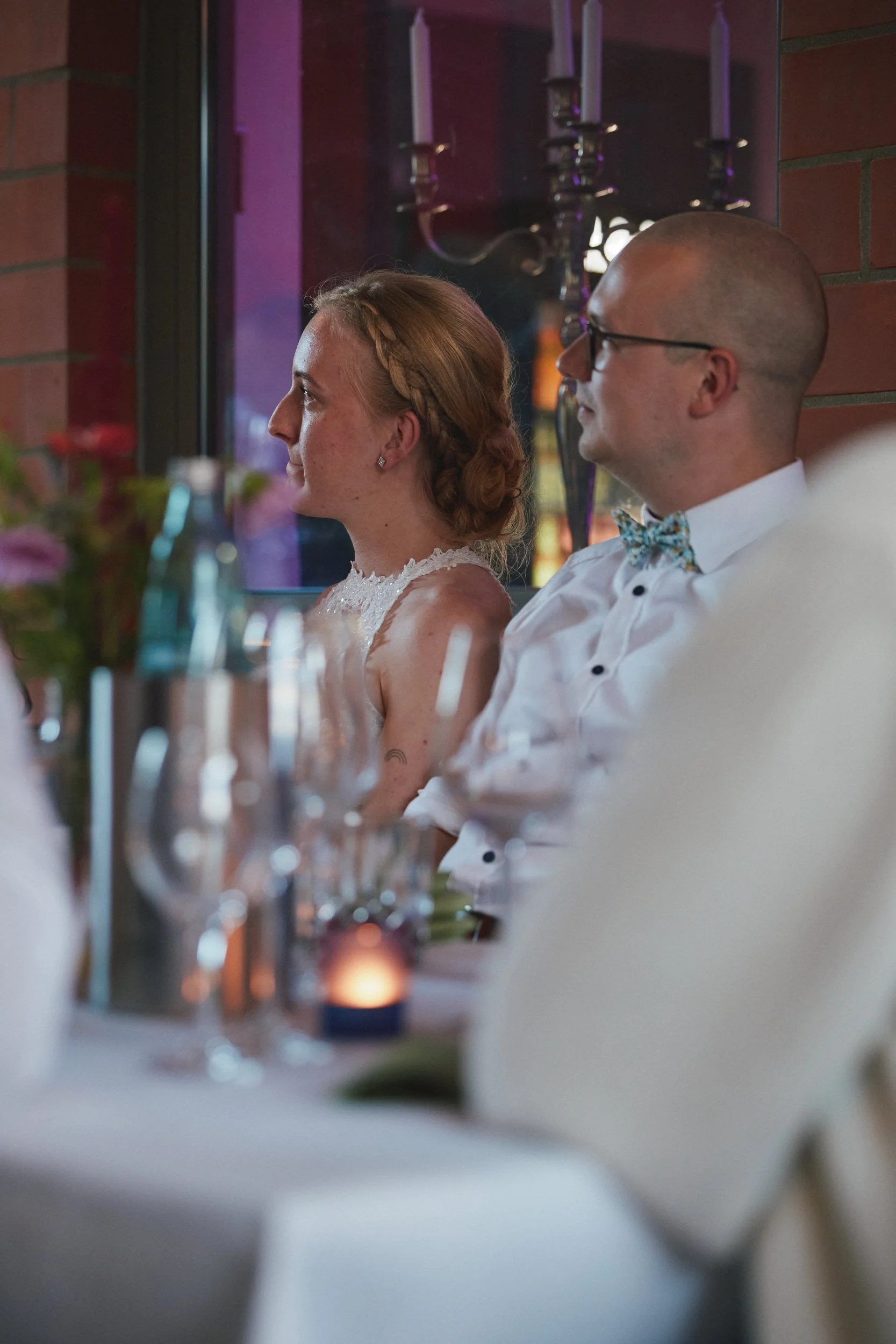 Ein Brautpaar sitzt ruhig und aufmerksam bei einer Hochzeit, im Vordergrund sind Dekorationen und Kerzen, im Hintergrund eine Kerzenleuchter. Die Szene ist festlich und intimer Atmosphäre.